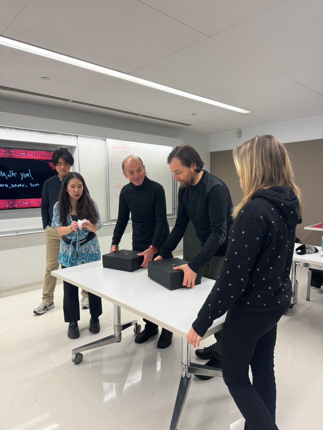 Group of people in a classroom gathered around a table with two black boxes, one man is opening a box, others are observing, whiteboard and a digital screen in the background.