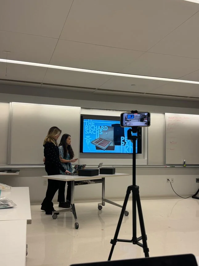 Two women standing at the front of a classroom presenting, with a large screen behind them showing text and images, a camera on a tripod recording the presentation, and a whiteboard on the right side of the room.