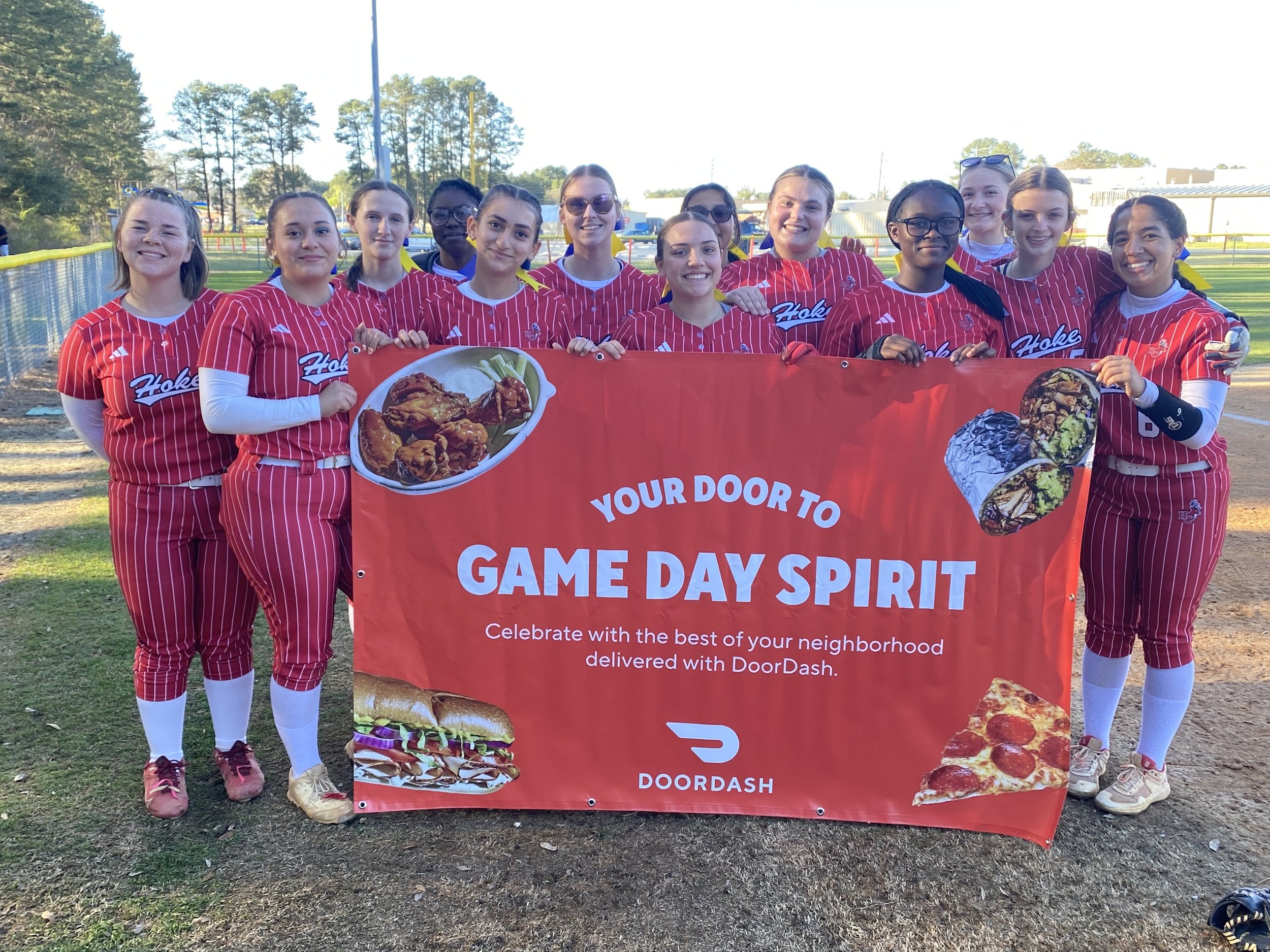 A group of softball players in red striped uniforms hold a red DoorDash banner promoting "Game Day Spirit" on a field.
