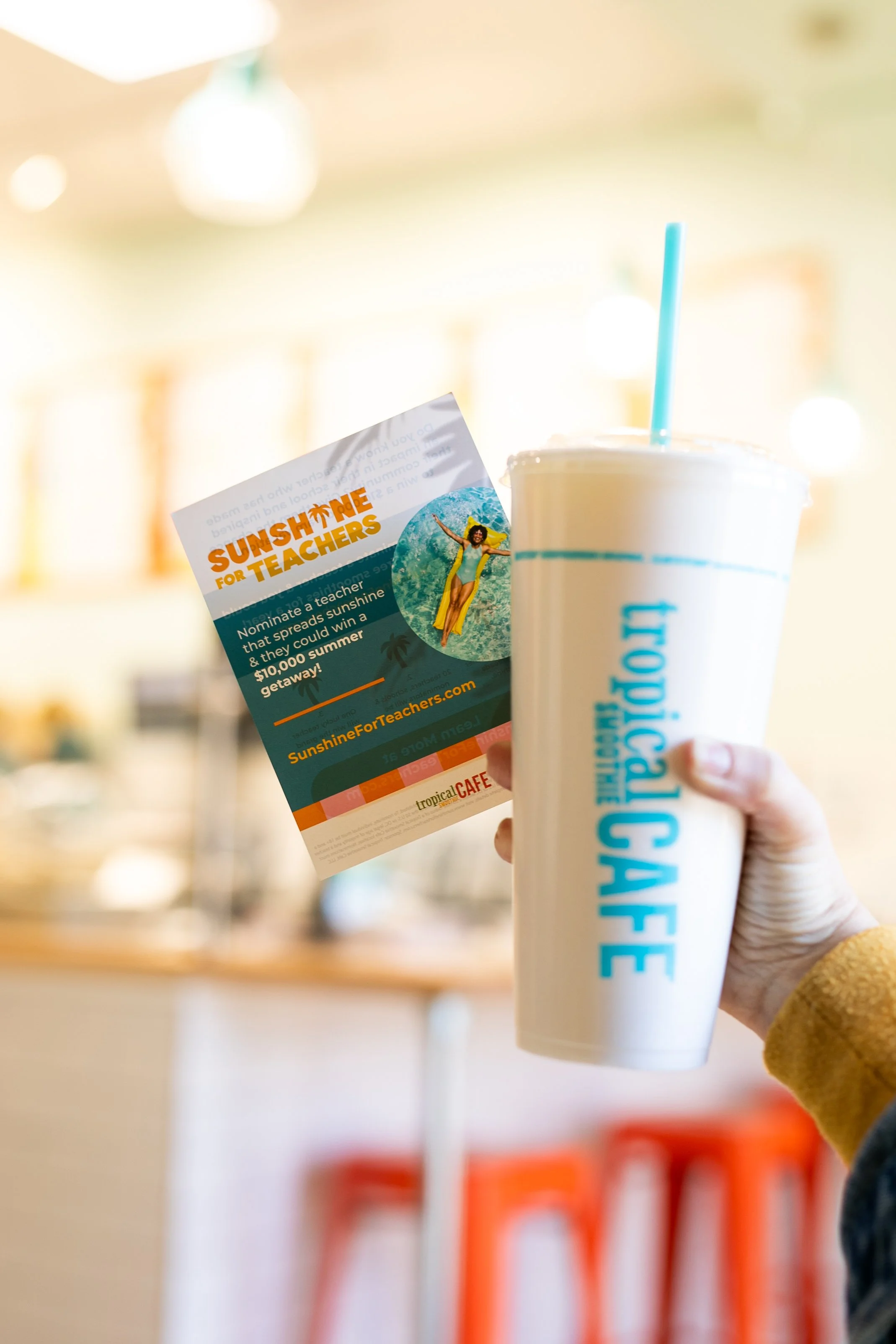 A person holding a large cup with the Tropical Smoothie Cafe logo and a promotional flyer for "Sunshine for Teachers" in a cafe setting.