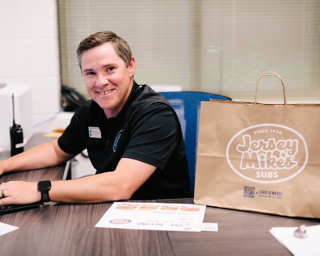 A person sitting at a desk next to a Jersey Mike's Subs paper bag, smiling at the camera. The desk has a computer, papers, and a walkie-talkie on it.