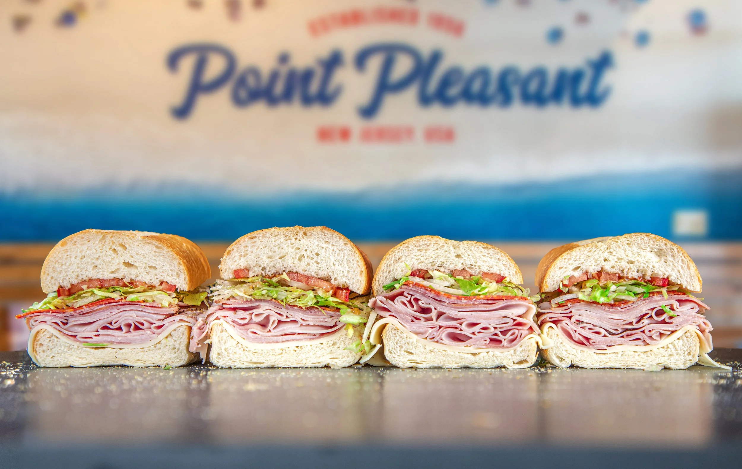 Four sub sandwiches with ham, lettuce, tomato, and cheese, placed in a row on a counter, with "Point Pleasant, New Jersey" sign in the background.