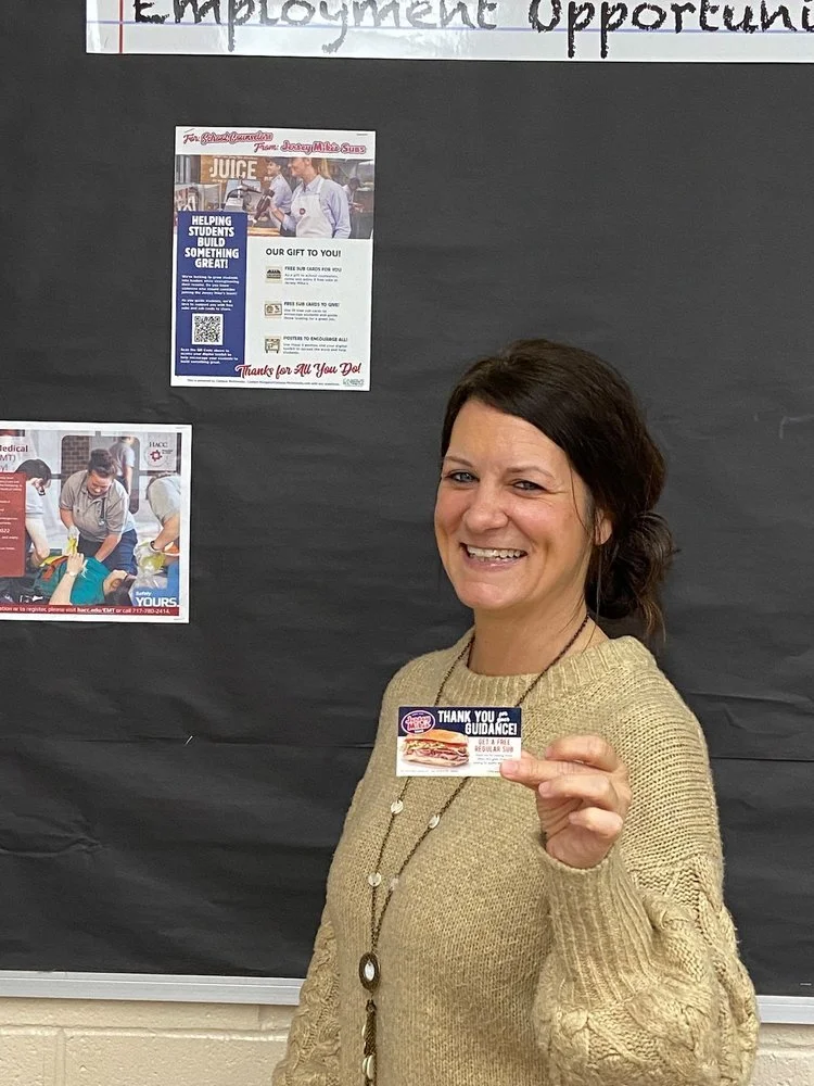 Woman in a beige sweater holding a gift card, standing in front of a bulletin board with flyers and the words 'Employment Opportunities' displayed.