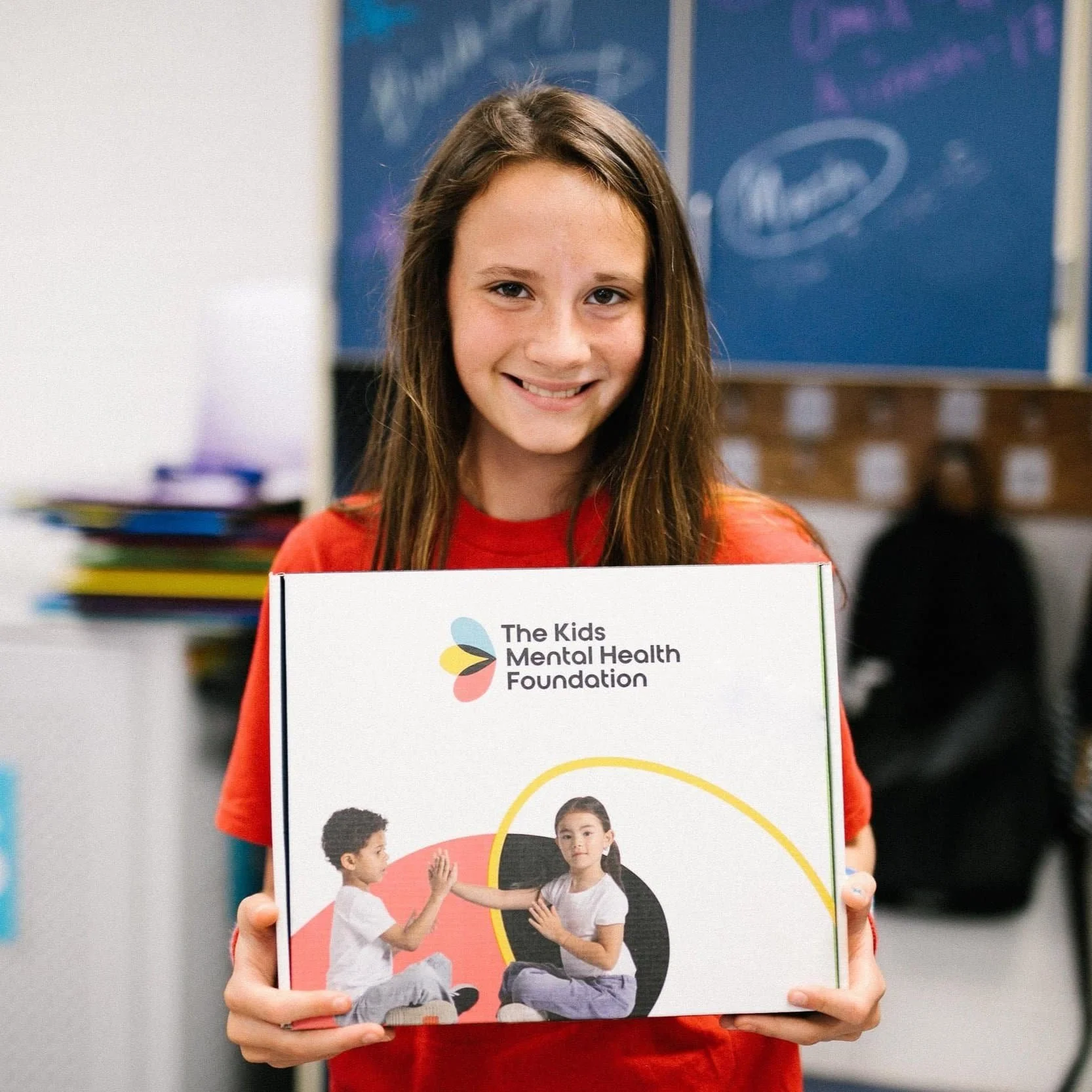 Girl holding a box labeled 'The Kids Mental Health Foundation' with an image of two children playing on it.