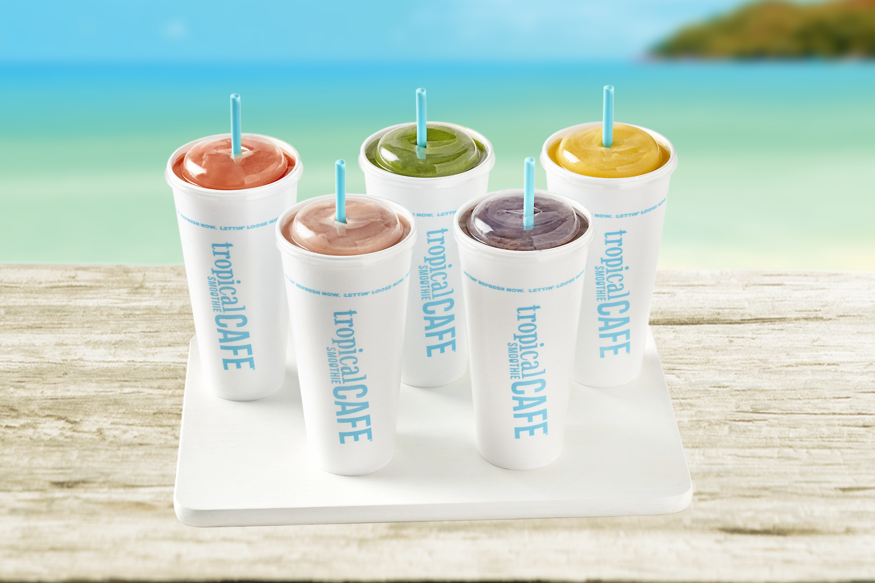 Five Tropical Smoothie Cafe cups with different smoothie colors on a table by the beach.
