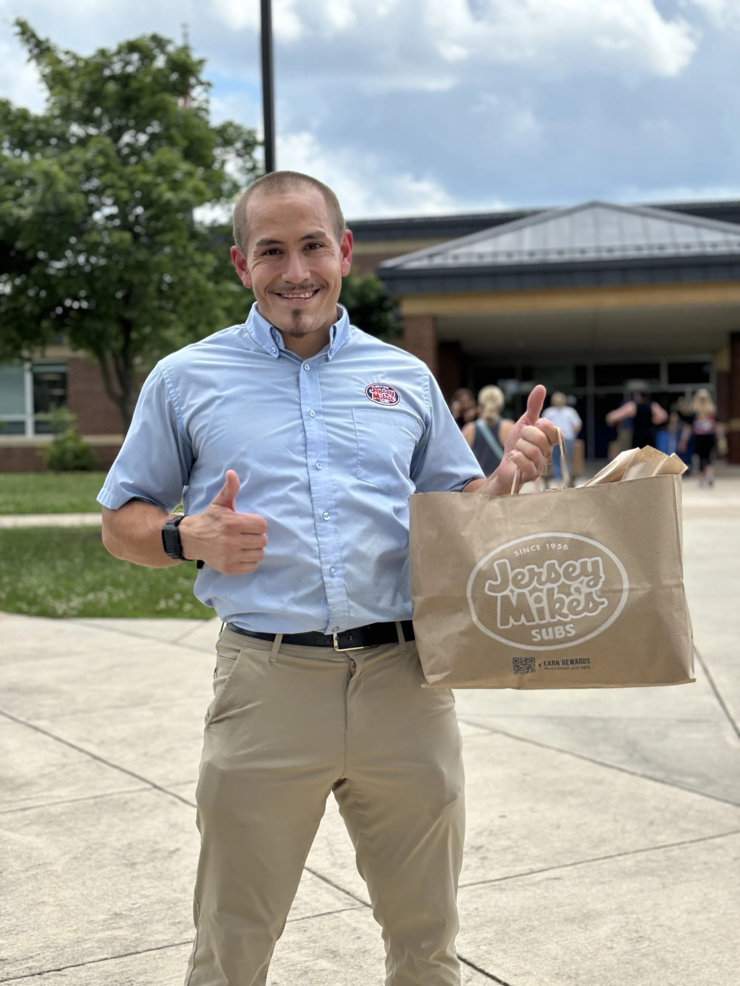 Person holding a Jersey Mike's Subs bag, standing outside a building on a sunny day.