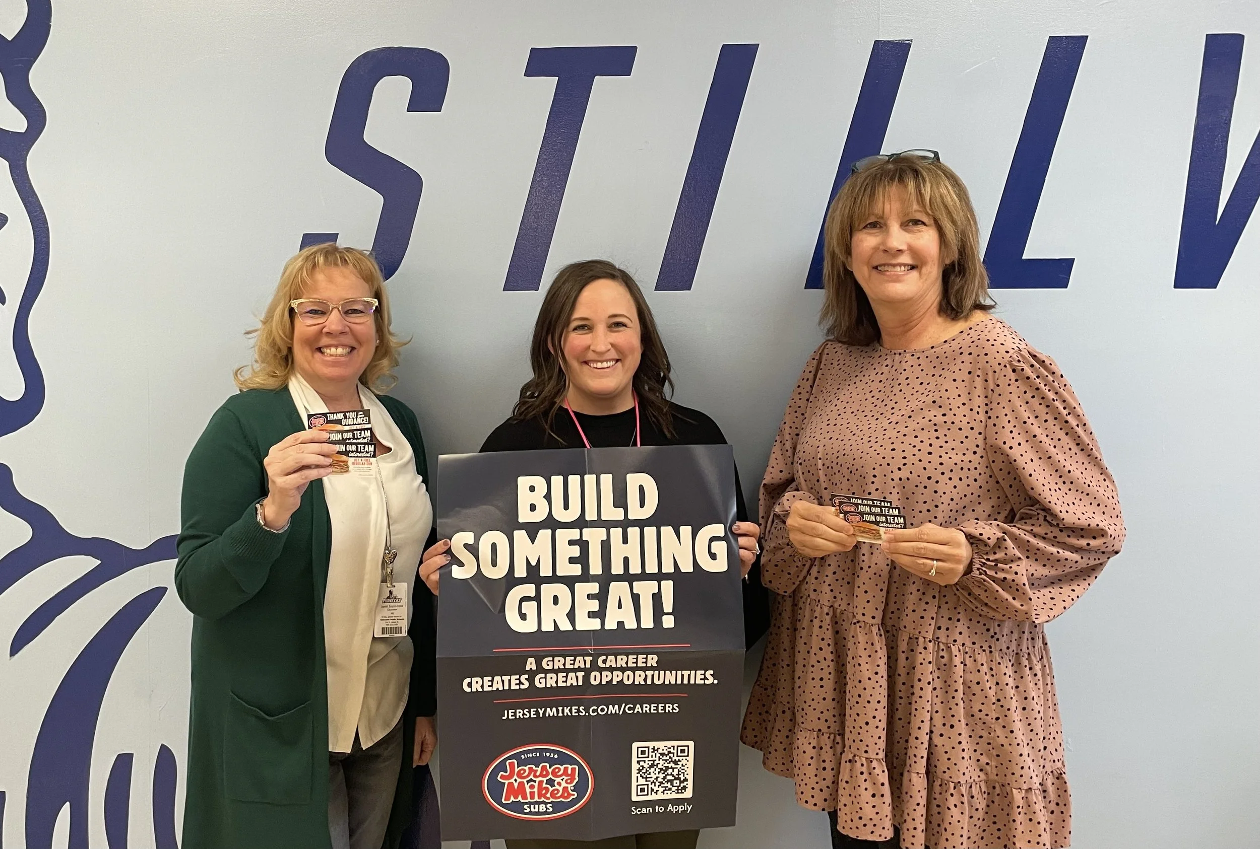 Three women smiling and holding a "Build Something Great!" sign promoting Jersey Mike's Subs careers.