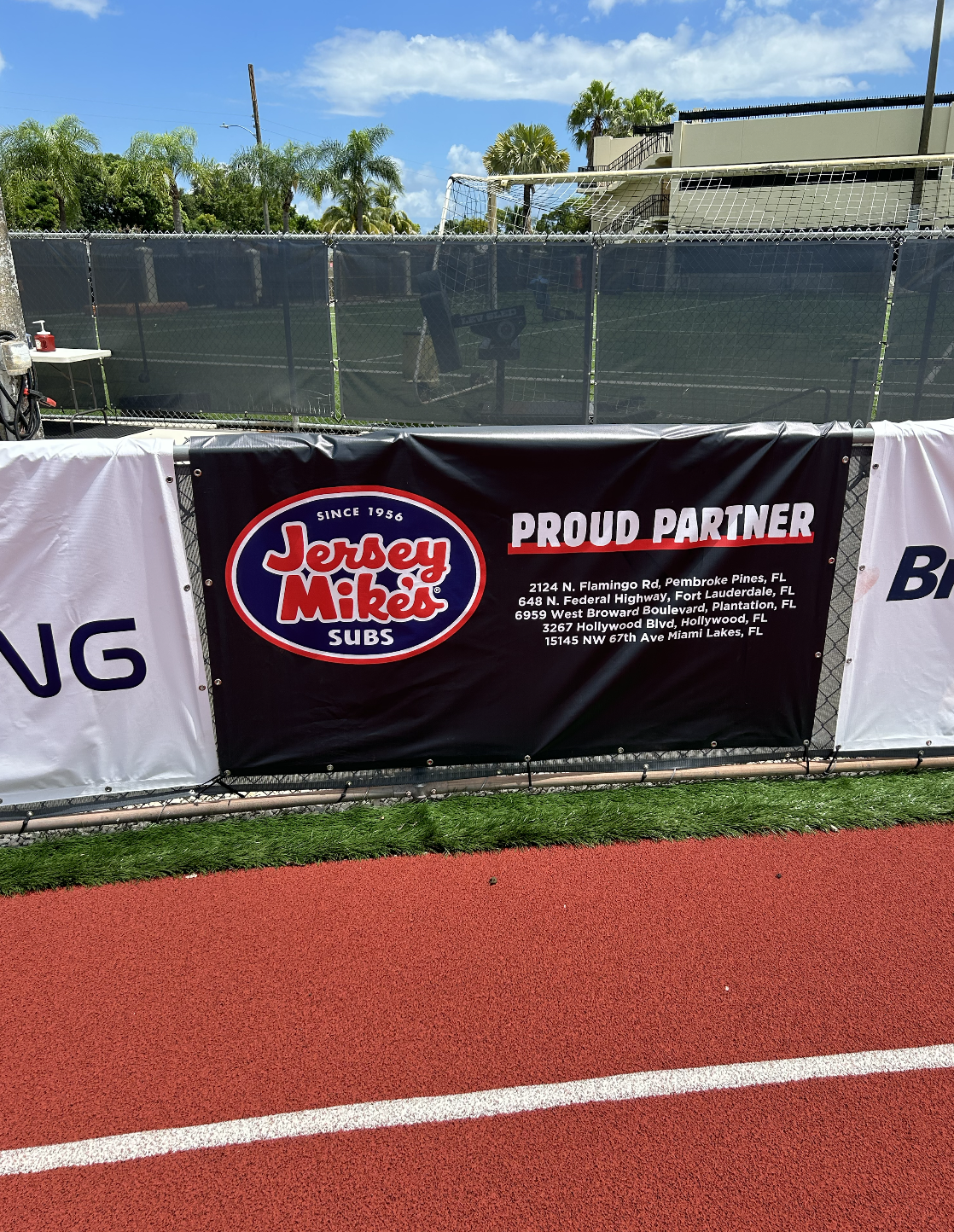 Banner advertising Jersey Mike's Subs as a proud partner, displayed on a football field fence. Includes multiple Florida addresses under the logo.
