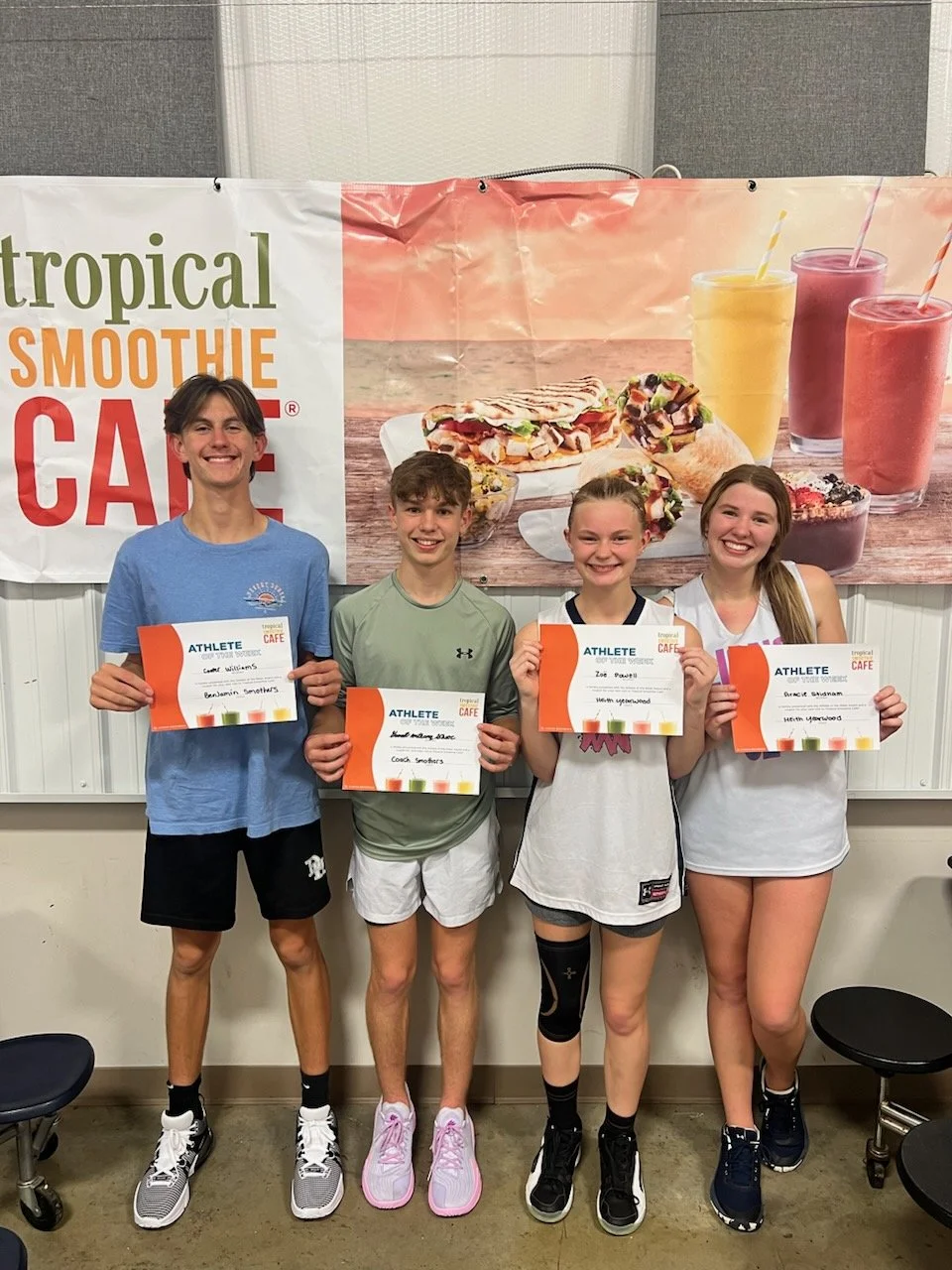 Four young athletes standing in front of a Tropical Smoothie Cafe banner, each holding a certificate. The banner displays images of smoothies and food. The athletes are smiling and wearing casual athletic clothing.