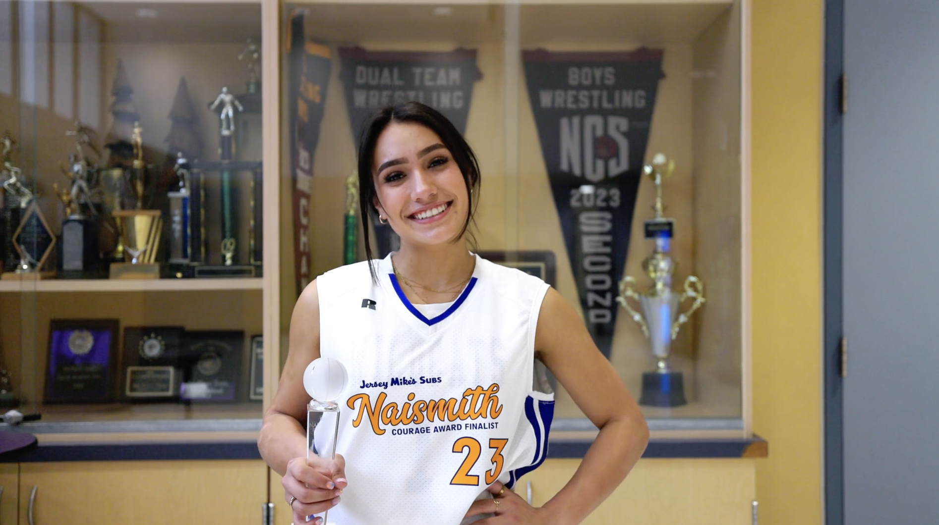 A female athlete in a white sports jersey holding a trophy, standing in front of a display case with awards and banners in the background.
