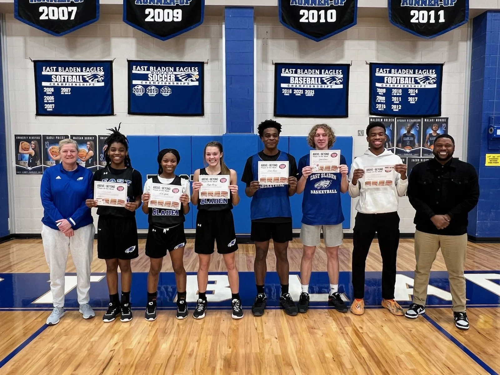 A group of seven people standing in a gymnasium displaying certificates, with "East Bladen Eagles" banners and sports achievements in the background. Four individuals in the middle are wearing basketball uniforms.