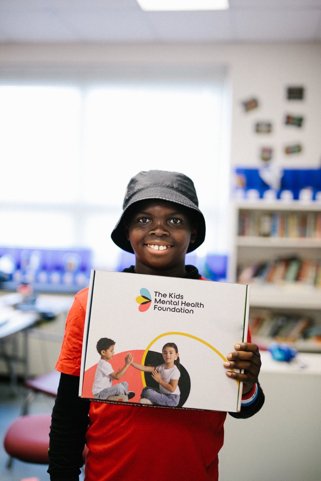Smiling child holding a box labeled 'The Kids Mental Health Foundation' in a classroom setting.