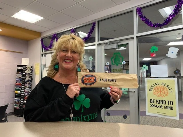 A woman smiling while holding a Jersey Mike's wrapped sandwich in an office decorated with festive elements and a mental health sign on the wall.