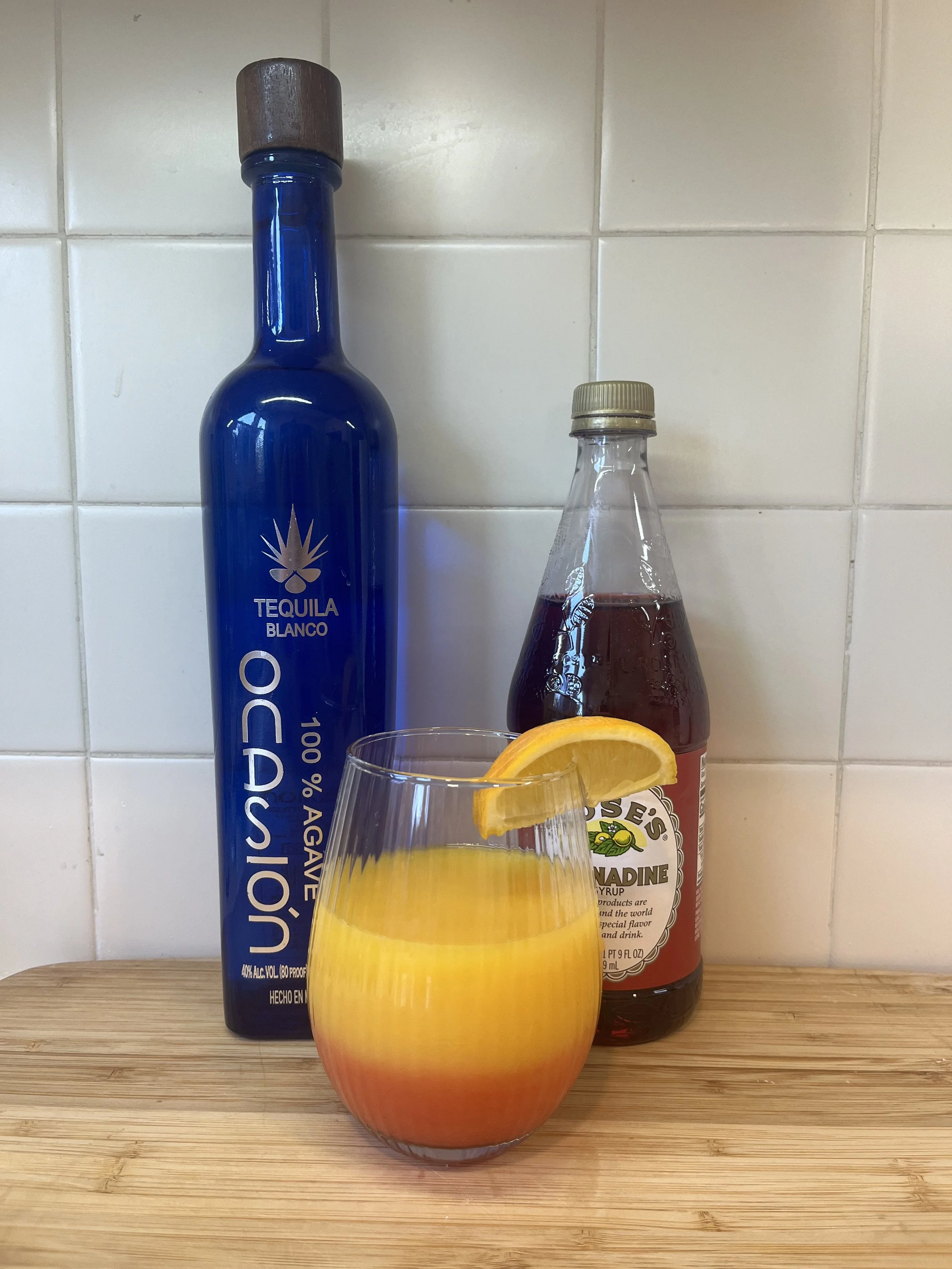 A glass of orange juice with a lemon slice on the rim, placed on a wooden surface. Behind it are a blue bottle of tequila and a smaller bottle of grenadine syrup, set against a white tiled wall.