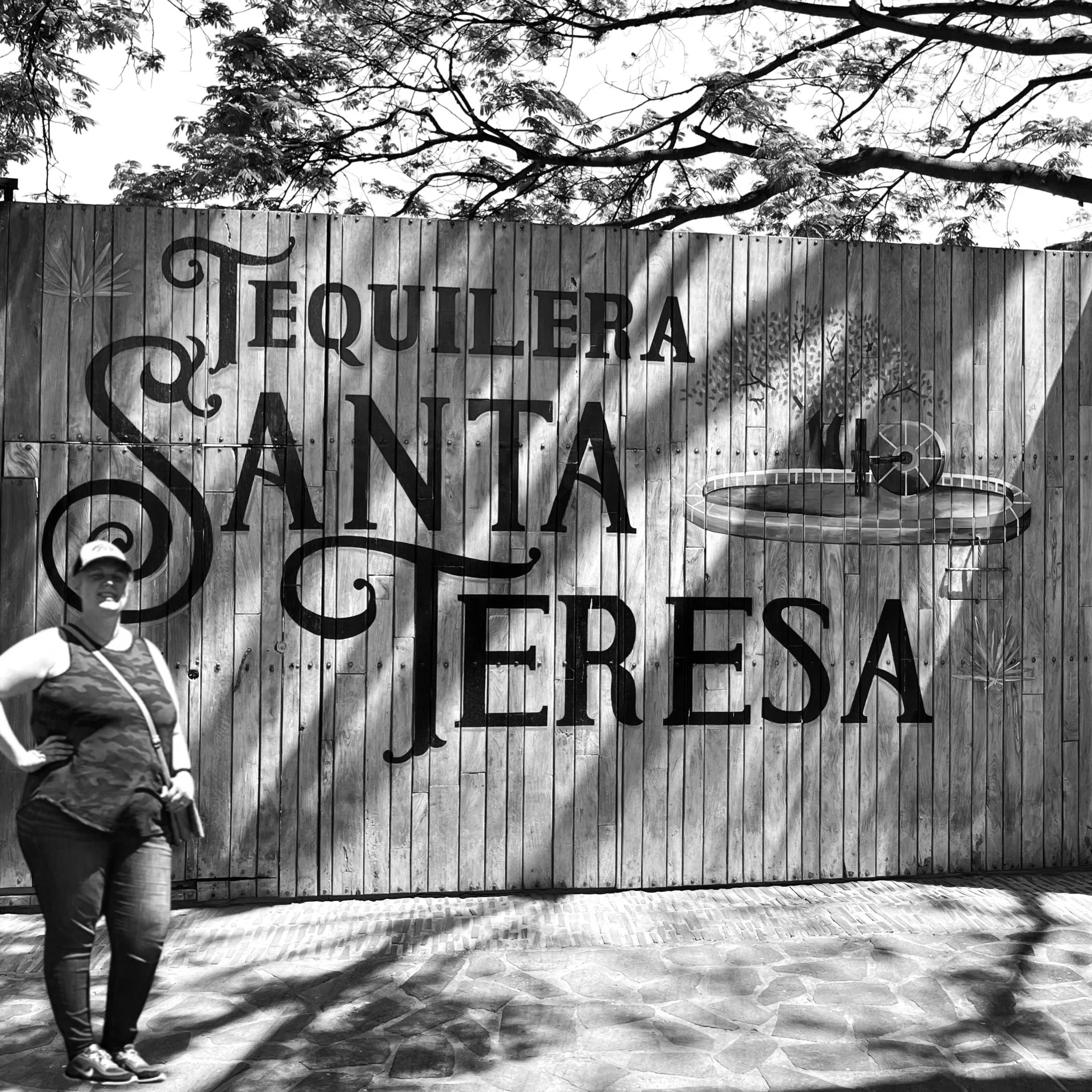 A woman standing in front of a wooden fence with the sign 'Tequileria Santa Teresa' painted on it. The sign also features a painted water wheel and some trees in the background.