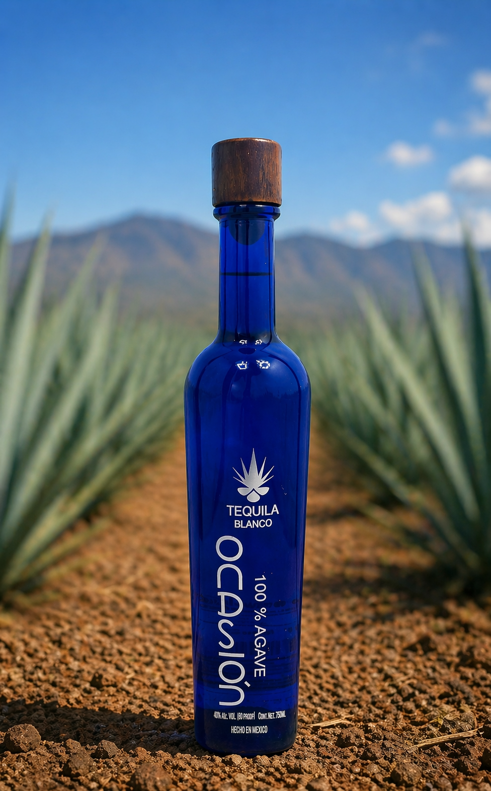 Blue bottle of tequila Blanco Ocasion with a wooden cap, standing in a field of agave plants with mountains and a blue sky in the background.