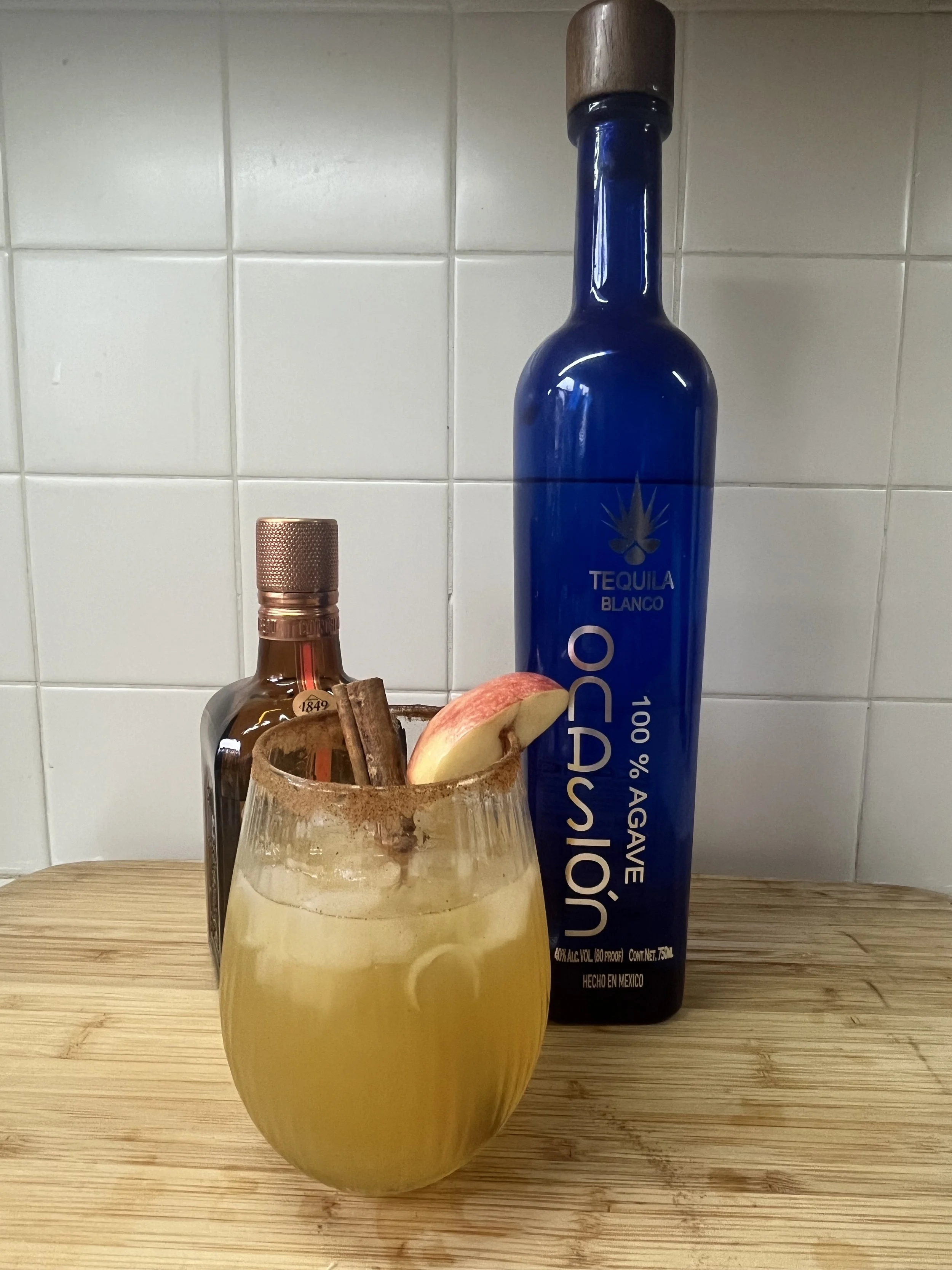 A blue bottle of tequila blanco, a glass with a cinnamon stick, apple slice, and ice, and a small bottle of alcohol on a wooden surface against a white tiled wall.