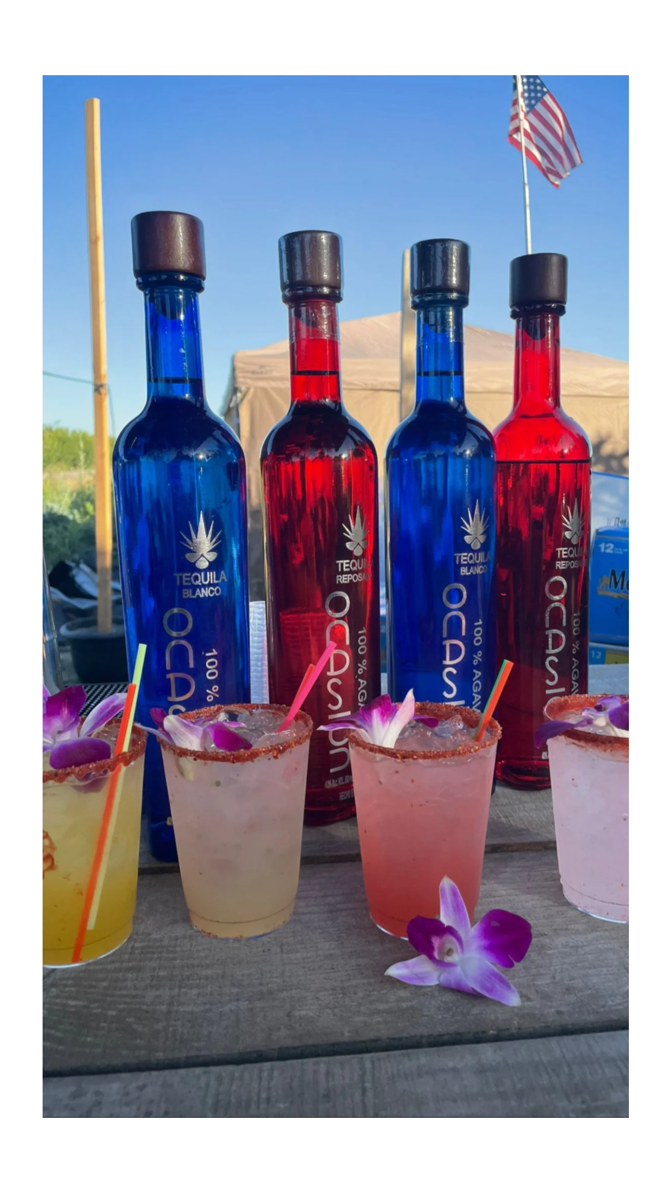 Four bottles of tequila, two with blue labels and two with red labels, are displayed behind four colorful cocktails garnished with orchid flowers and lined with colorful straws on a wooden table. The background shows a blue sky and a tent, with an American flag flying.