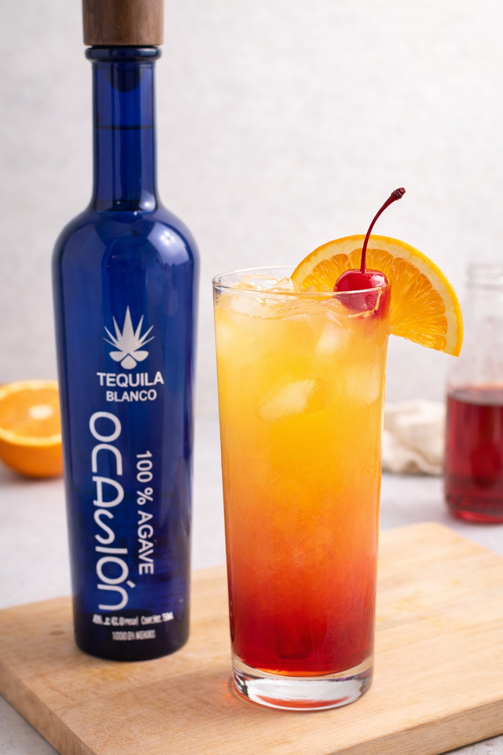 A glass of orange and red layered drink with an orange lemon slice on the rim, placed on a wooden surface in front of two bottles, one blue tequila bottle and one brown soda bottle, against a white tiled wall background.