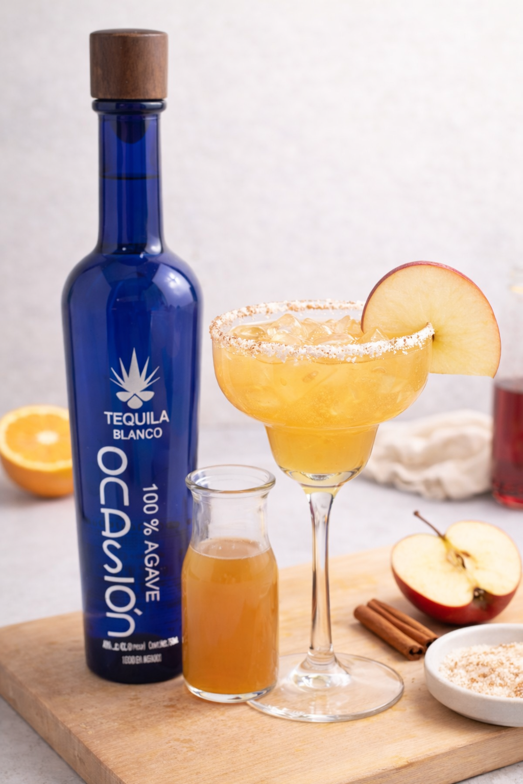 A glass of apple and cinnamon cocktail garnished with an apple slice and a cinnamon stick, with a bottle of tequila blanco and a smaller brown bottle behind it on a wooden surface against a white tiled wall.