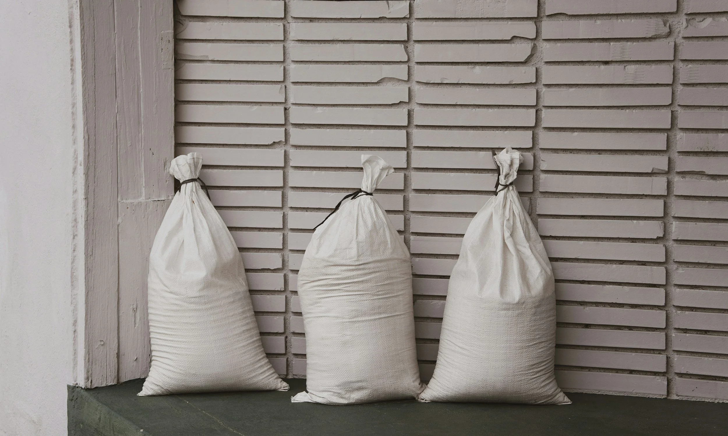 Three white sacks filled with an unknown material, tied at the top with black ties, leaning against a white textured brick wall on a green concrete surface.
