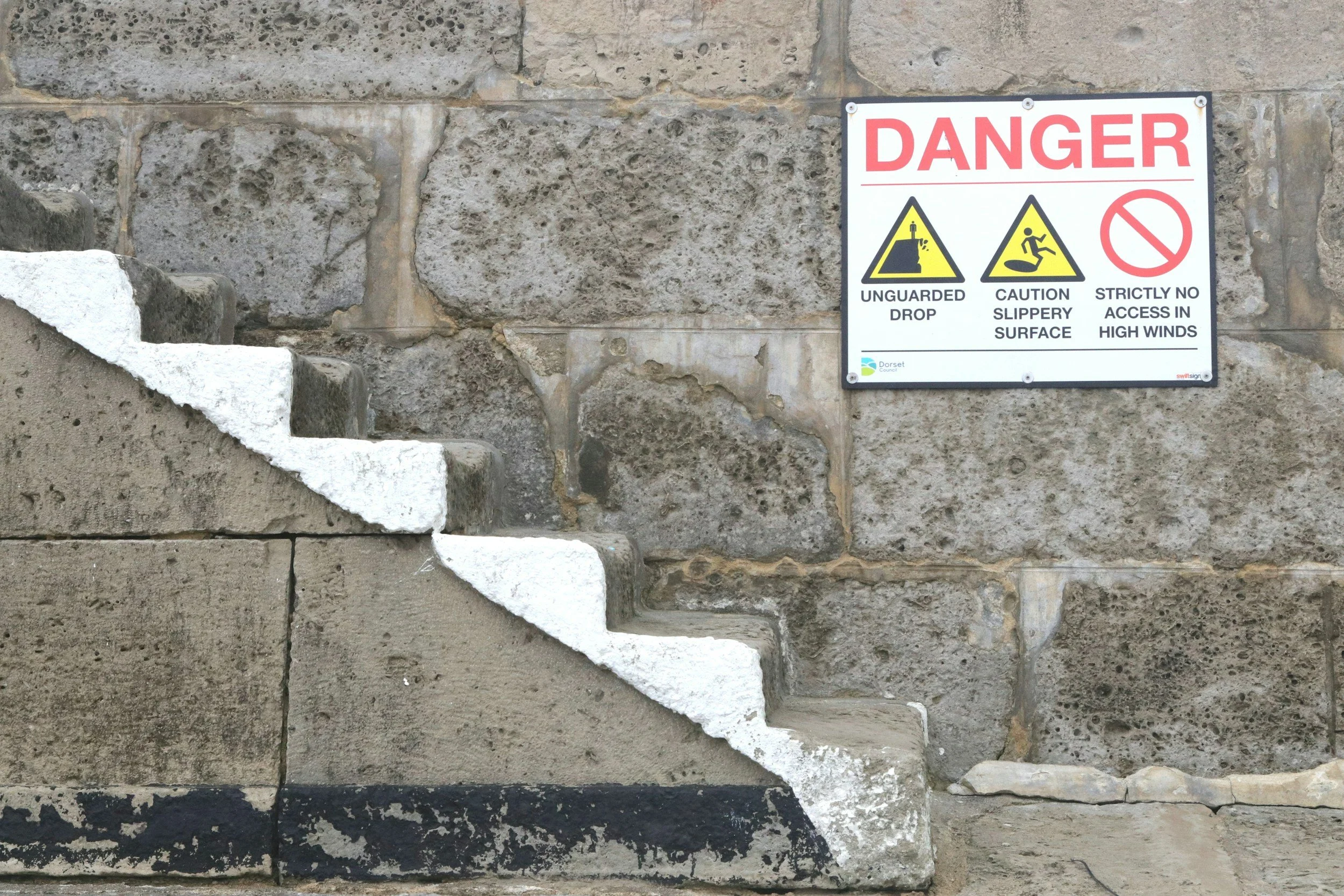 A stone staircase with a white painted edge and a warning sign on a stone wall. The sign says 'DANGER' in large red letters and indicates risks of unguarded drop, slippery surface, and no access in high winds.