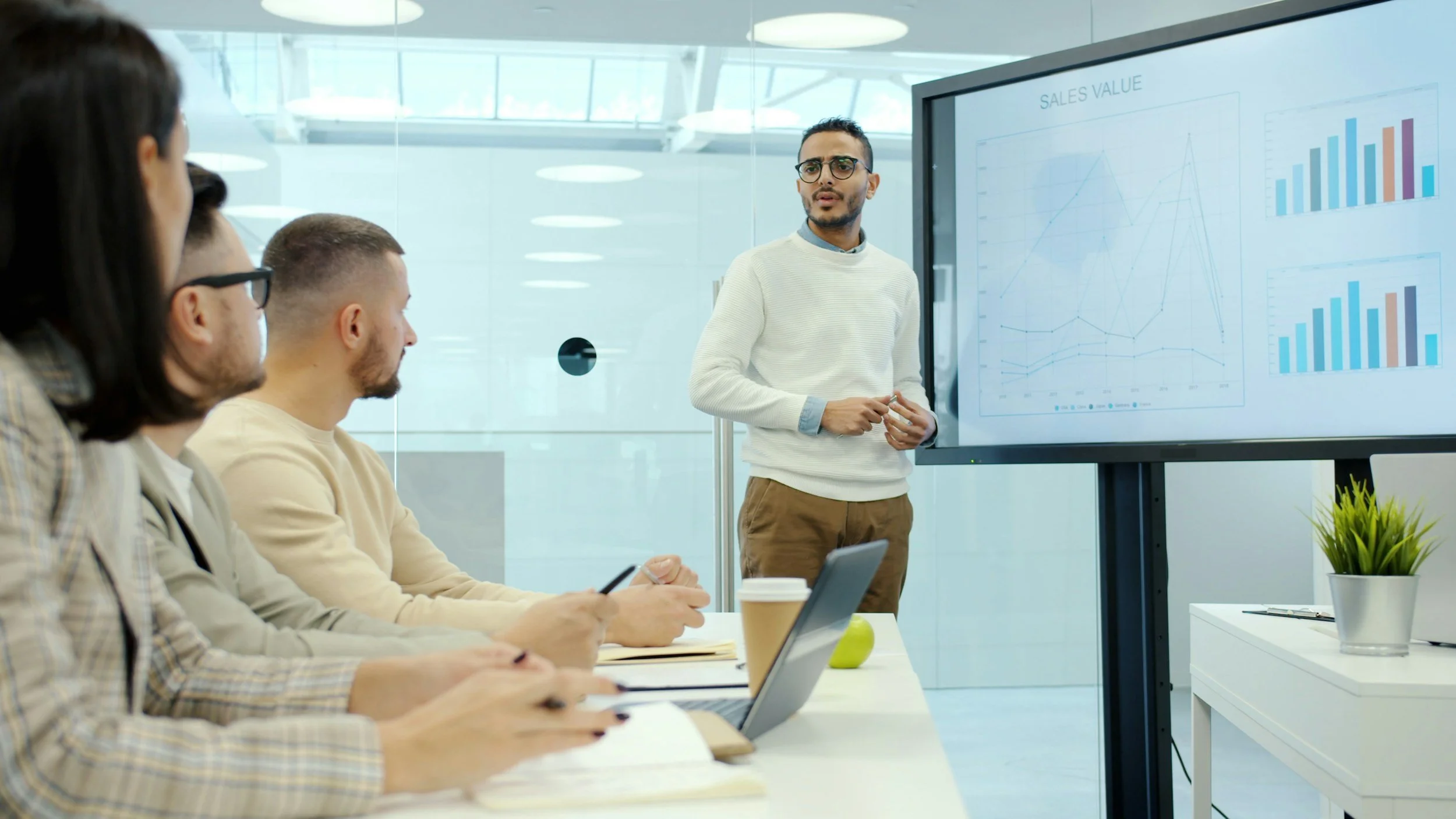 Business meeting with individuals seated at a table, listening to a presenter standing next to a large screen displaying sales data graphs.