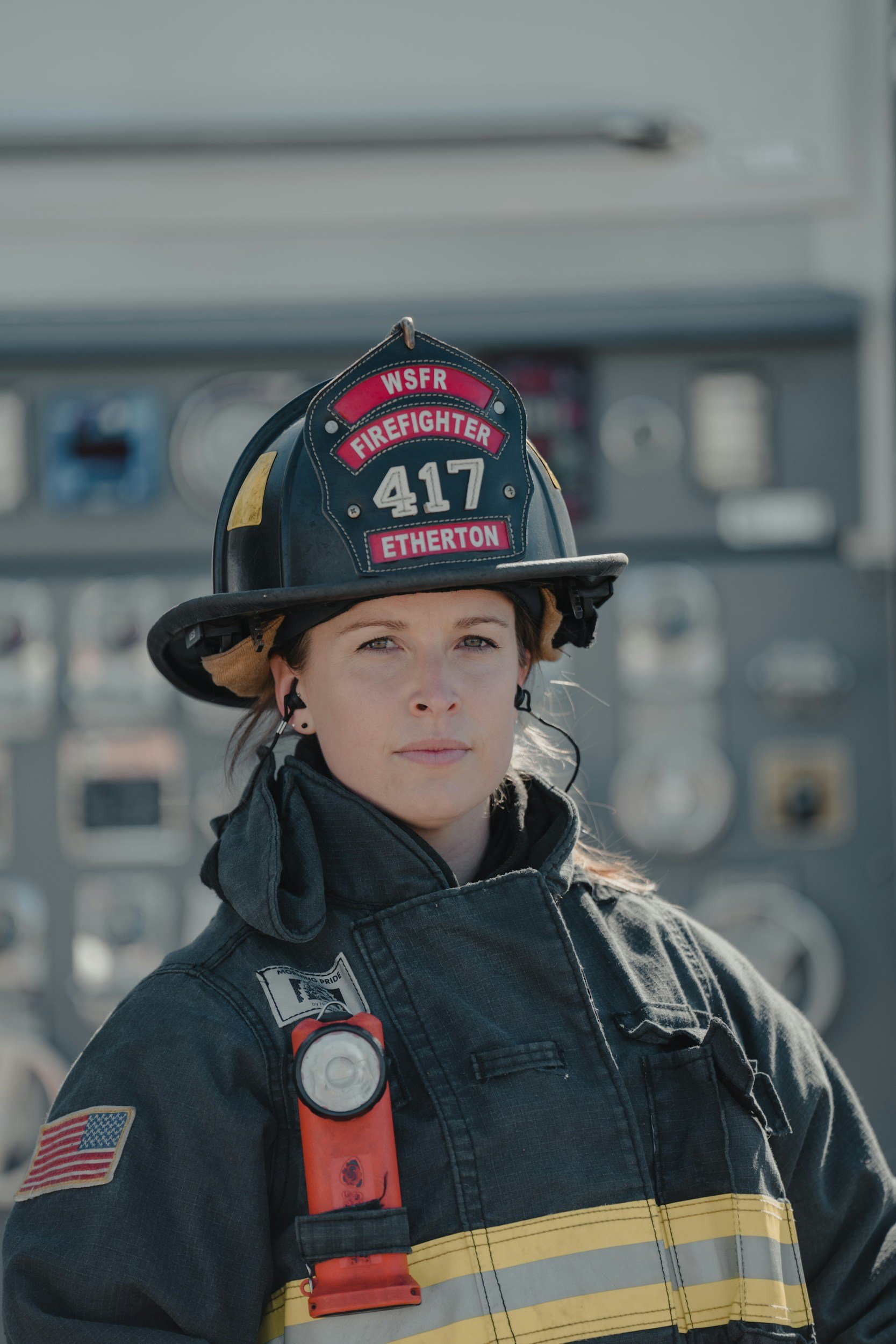 A young female firefighter in uniform and helmet looking at the camera, with a fire station panel in the background.