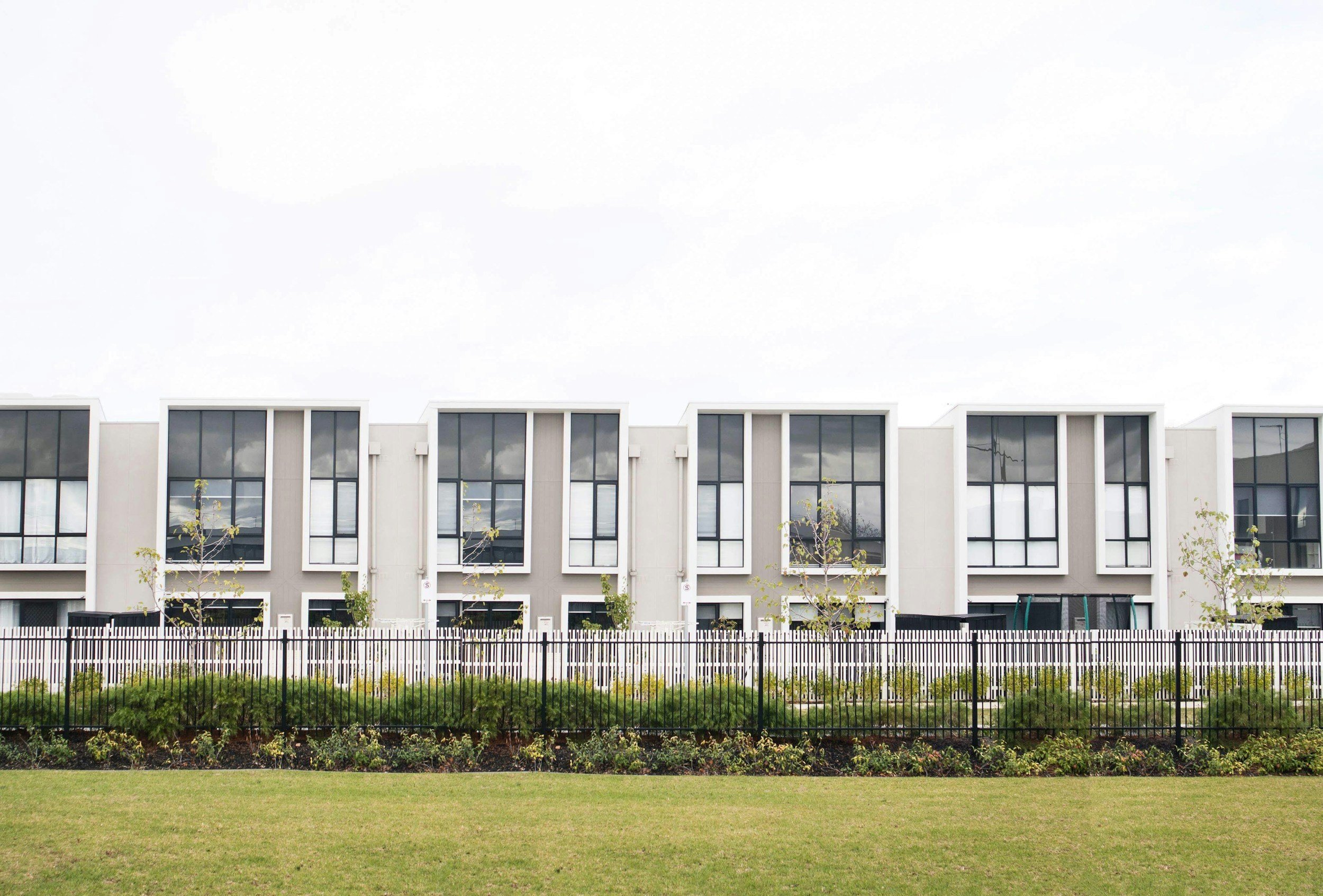 Modern residential building with large windows, small trees, and a black metal fence in front, under a cloudy sky.