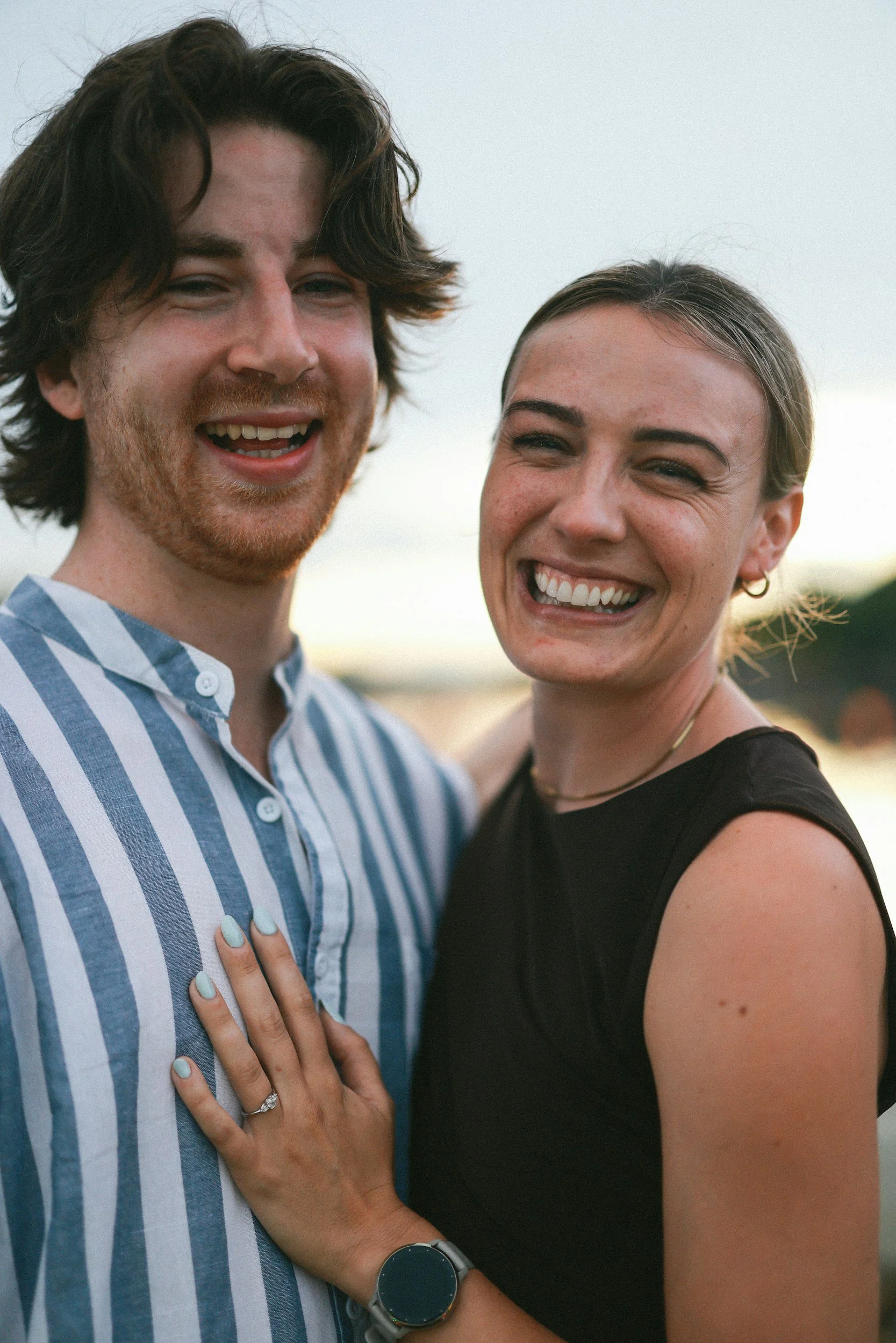 A happy couple smiling outdoors, with the woman hand resting on the man's chest, showing an engagement ring.
