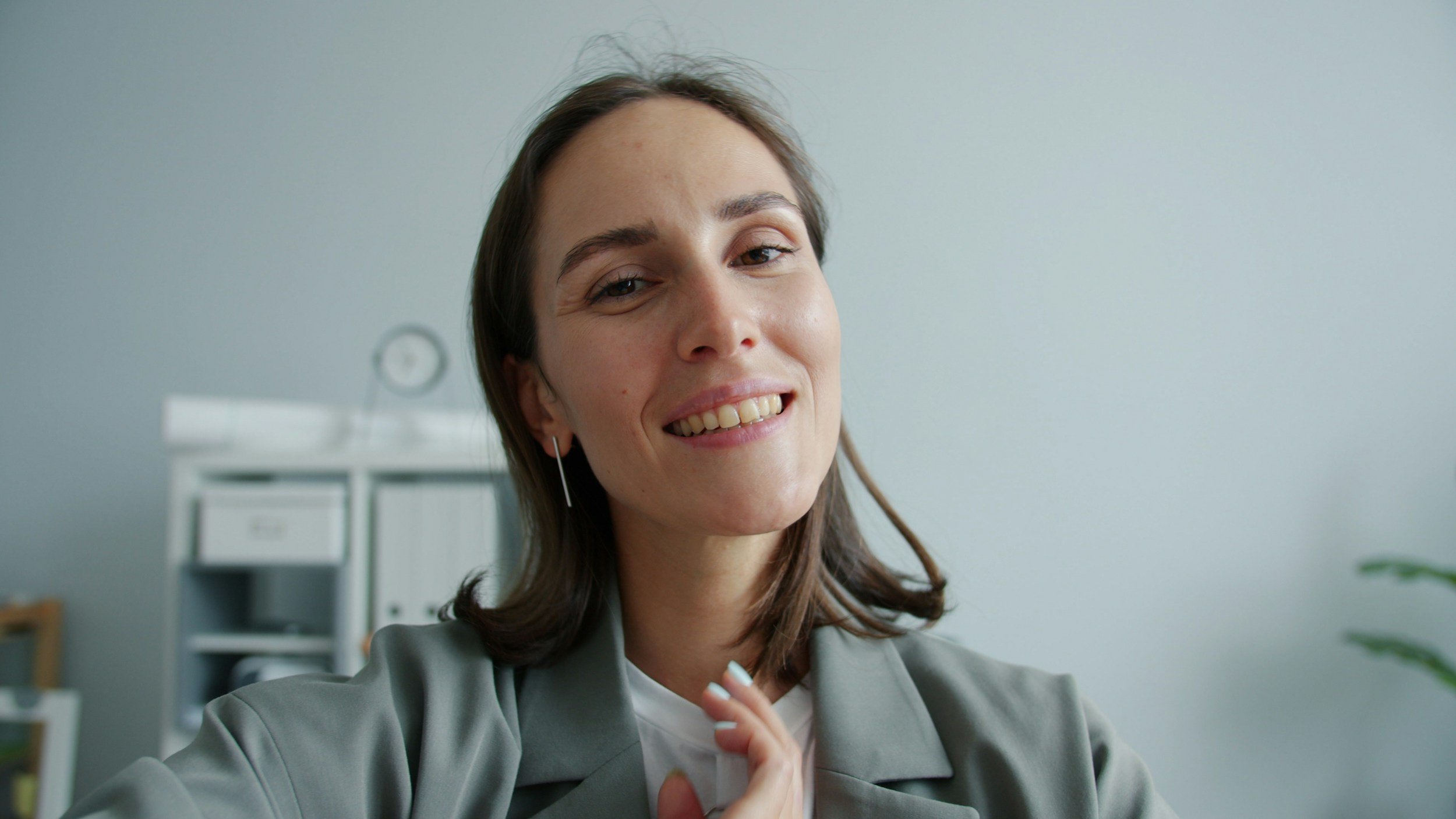 Close-up of a smiling woman with shoulder-length brown hair wearing a gray blazer, in a bright office space with a white cabinet and a clock in the background.