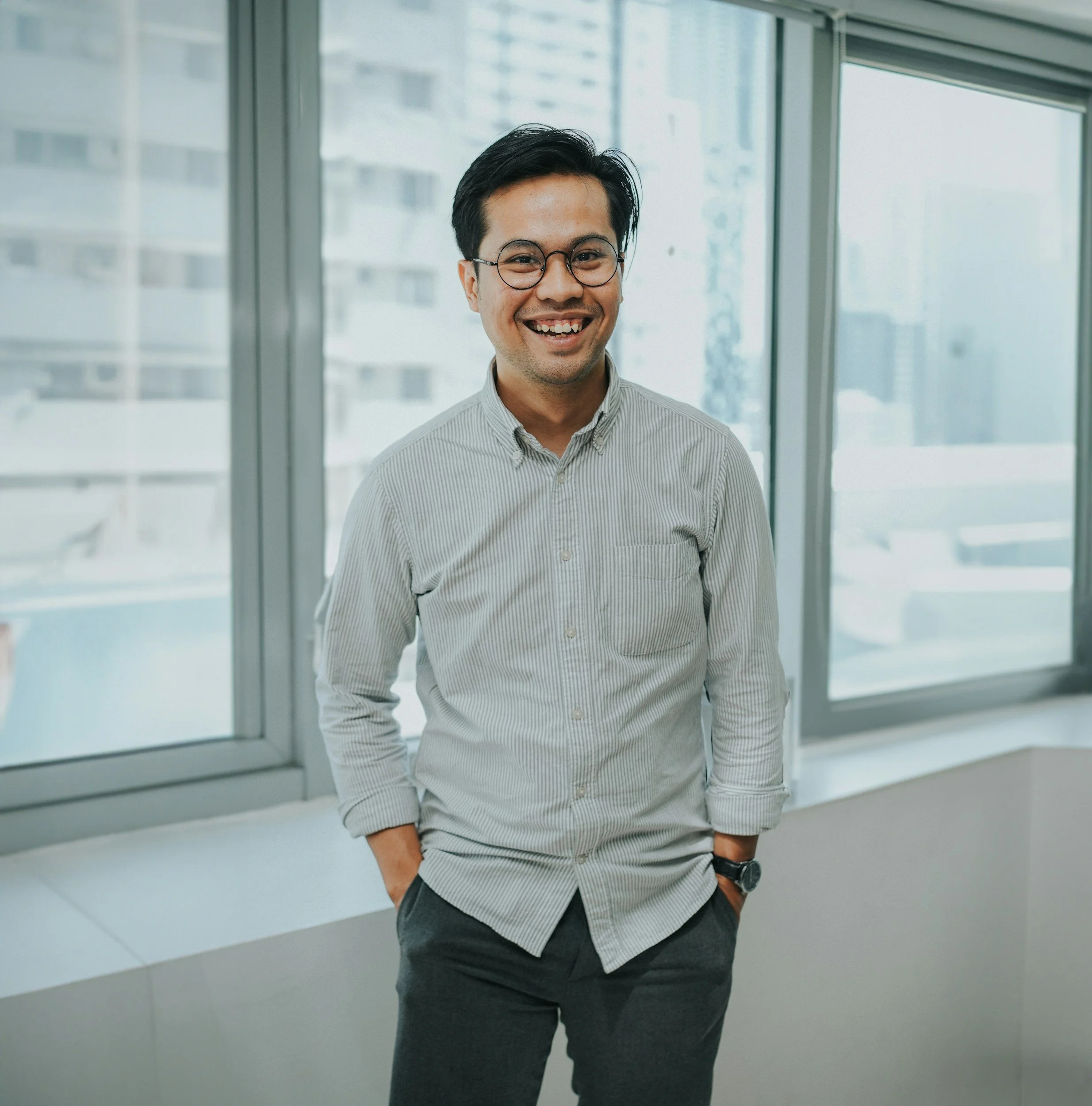 A smiling man with black hair, glasses, wearing a light gray striped button-up shirt with sleeves rolled up, dark gray pants, and a watch, standing in front of large windows with a cityscape background.