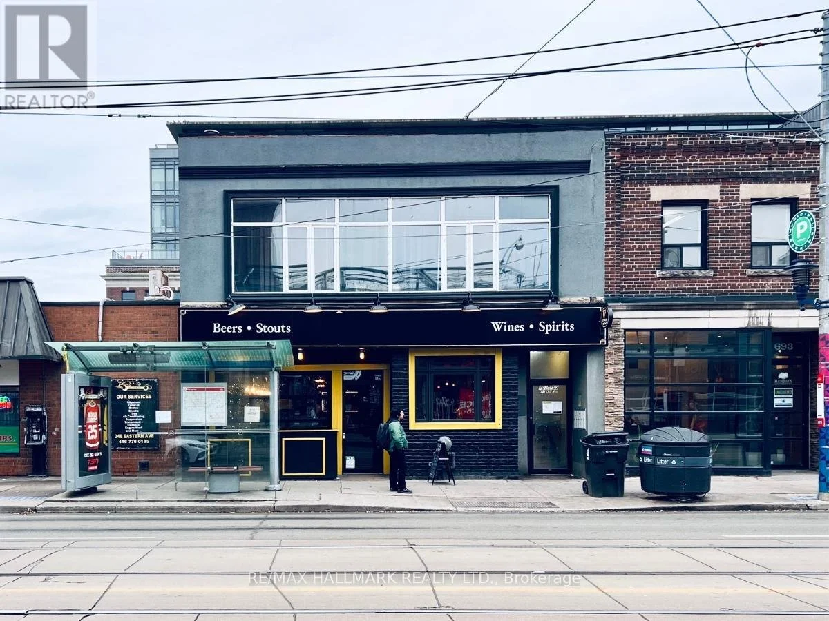 A storefront with a black awning displaying 'Beers • Stouts' on the left side and 'Wines • Spirits' on the right side, large windows, a sidewalk with a person standing, a bus stop shelter, trash bins, and a building with brick and gray exterior behind.