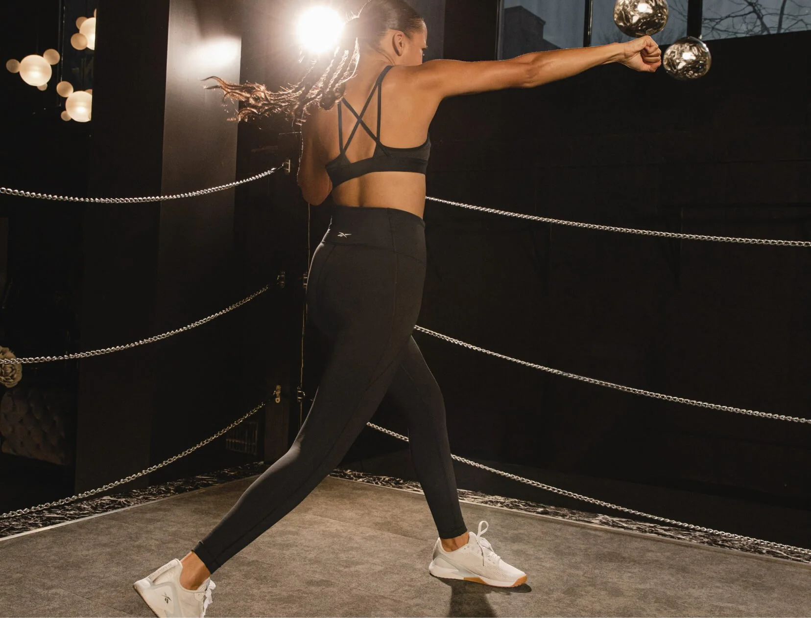 A woman in black athletic wear and white sneakers stretching in a gym with black walls and chain railings, with sunlight shining from behind her.