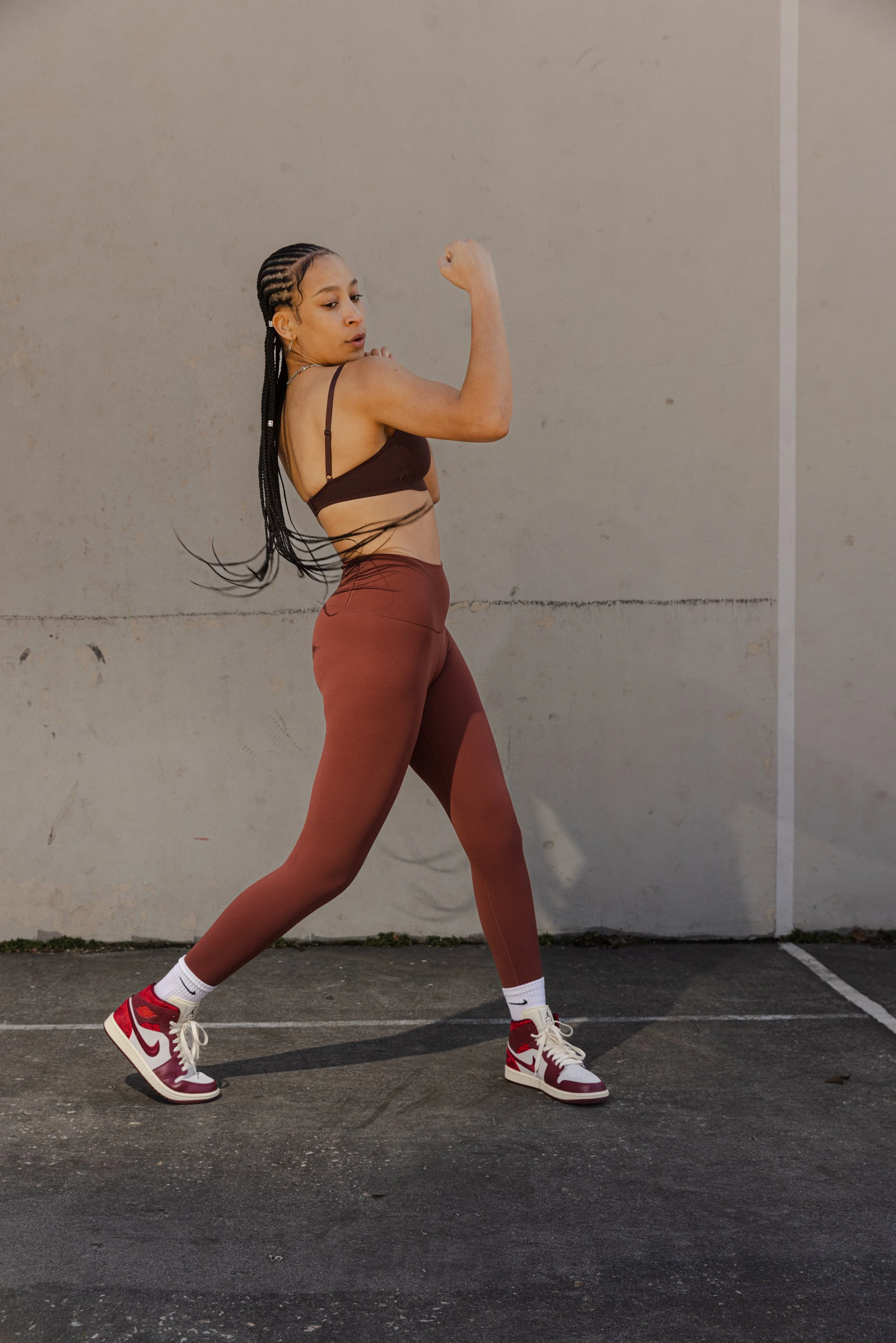 A young woman in athletic wear standing outdoors against a plain wall, flexing her bicep and looking at her arm.