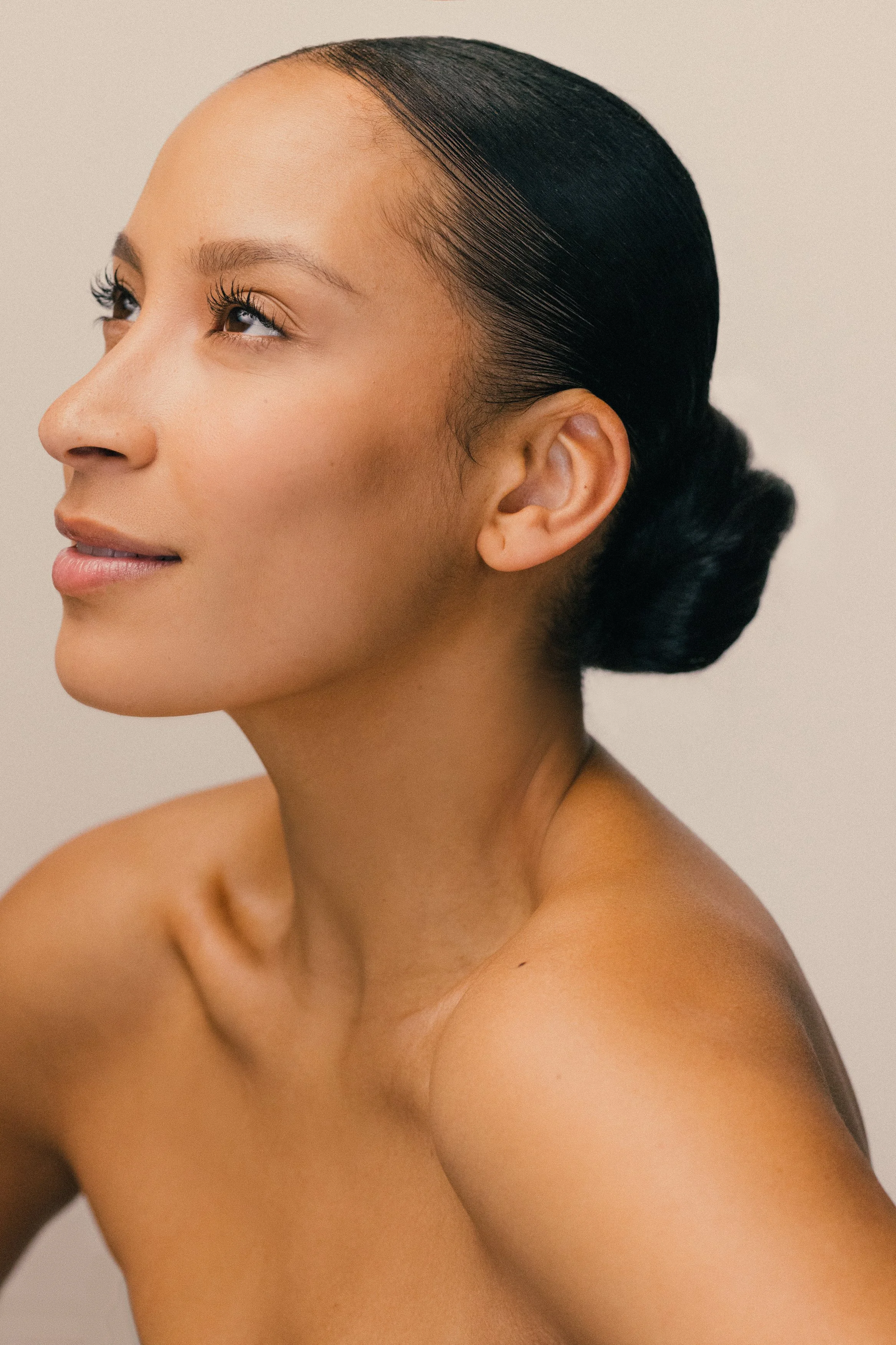 Close-up of a woman with black hair styled in a low bun, looking to the left, with smooth skin and a neutral expression.