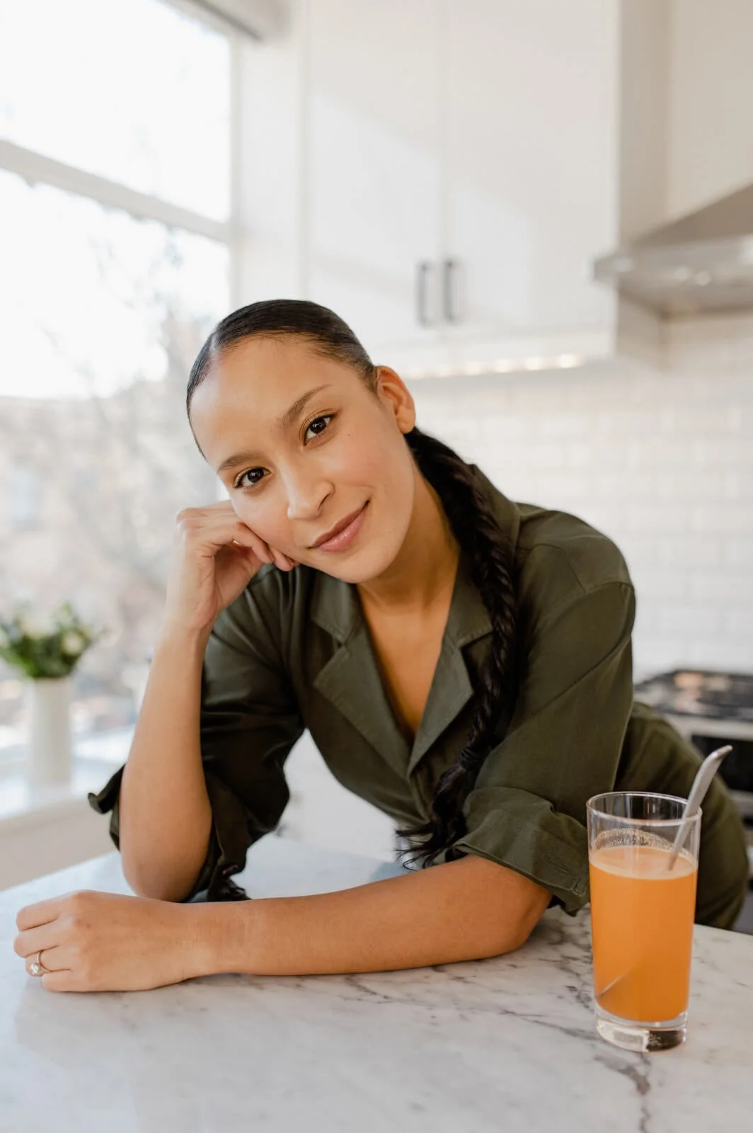 A woman with long dark hair in a braid leaning on her hand at a kitchen counter, with a glass of orange juice and a spoon, smiling at the camera.
