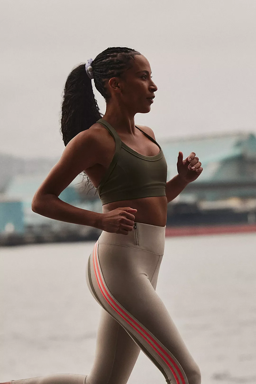 A woman running outdoors, wearing a green sports bra and beige leggings with pink and white stripes down the side.