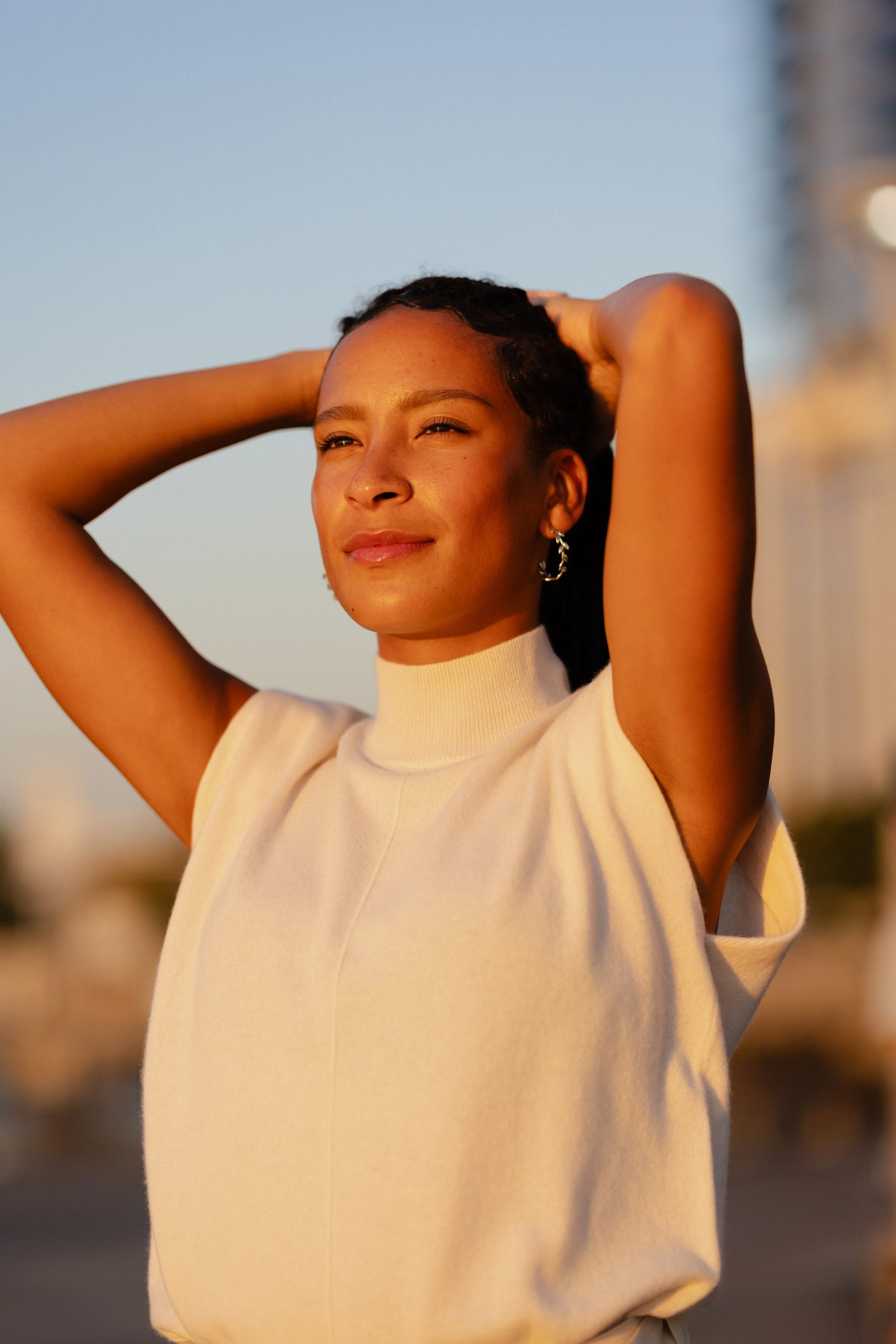 A woman with dark hair styled in waves, wearing a beige top and silver hoop earrings, standing outdoors during sunset with her hands behind her head, eyes closed, and a peaceful expression.