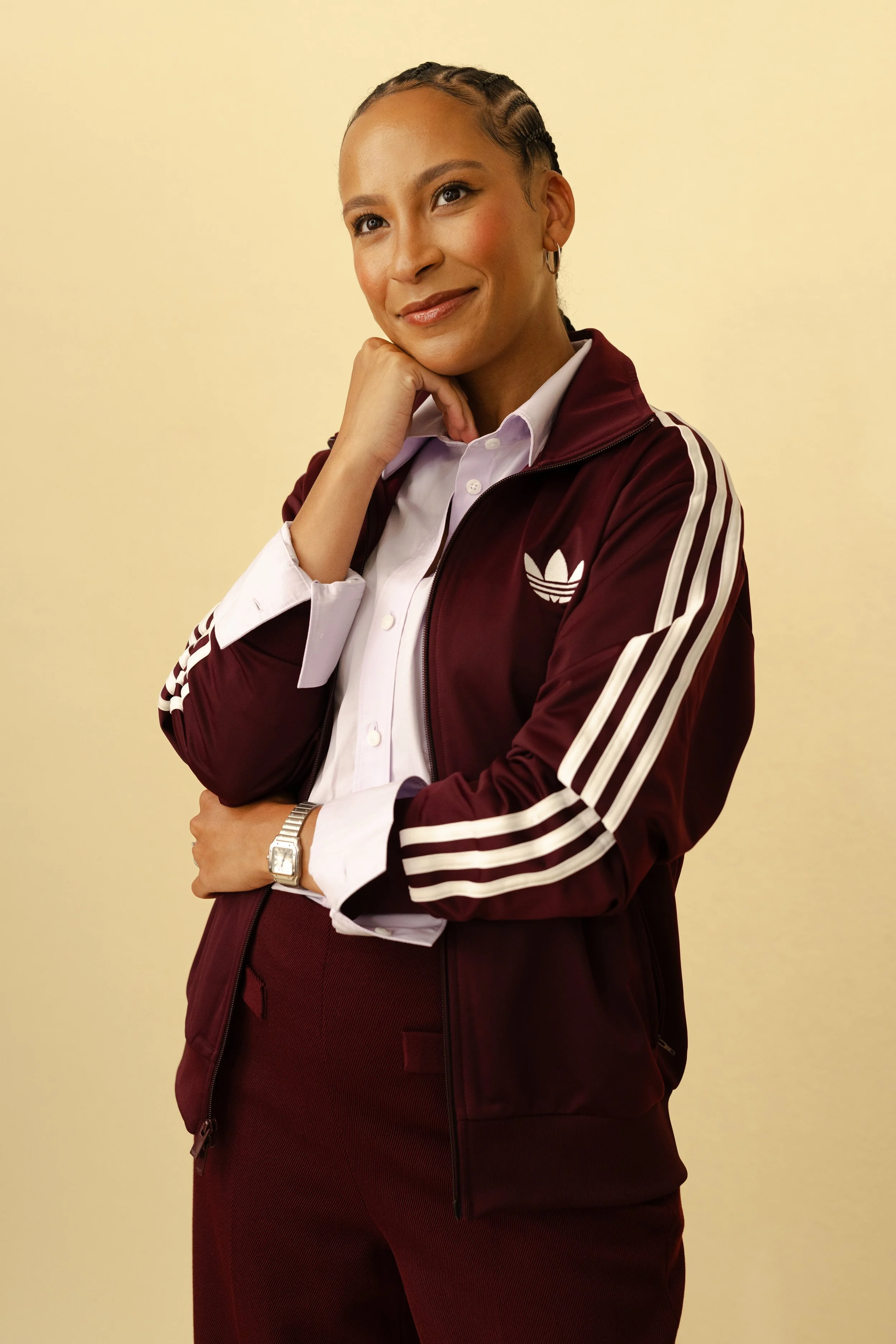 A woman with braided hair, wearing a white shirt and a maroon Adidas tracksuit jacket, posing with her hand on her chin against a light yellow background.