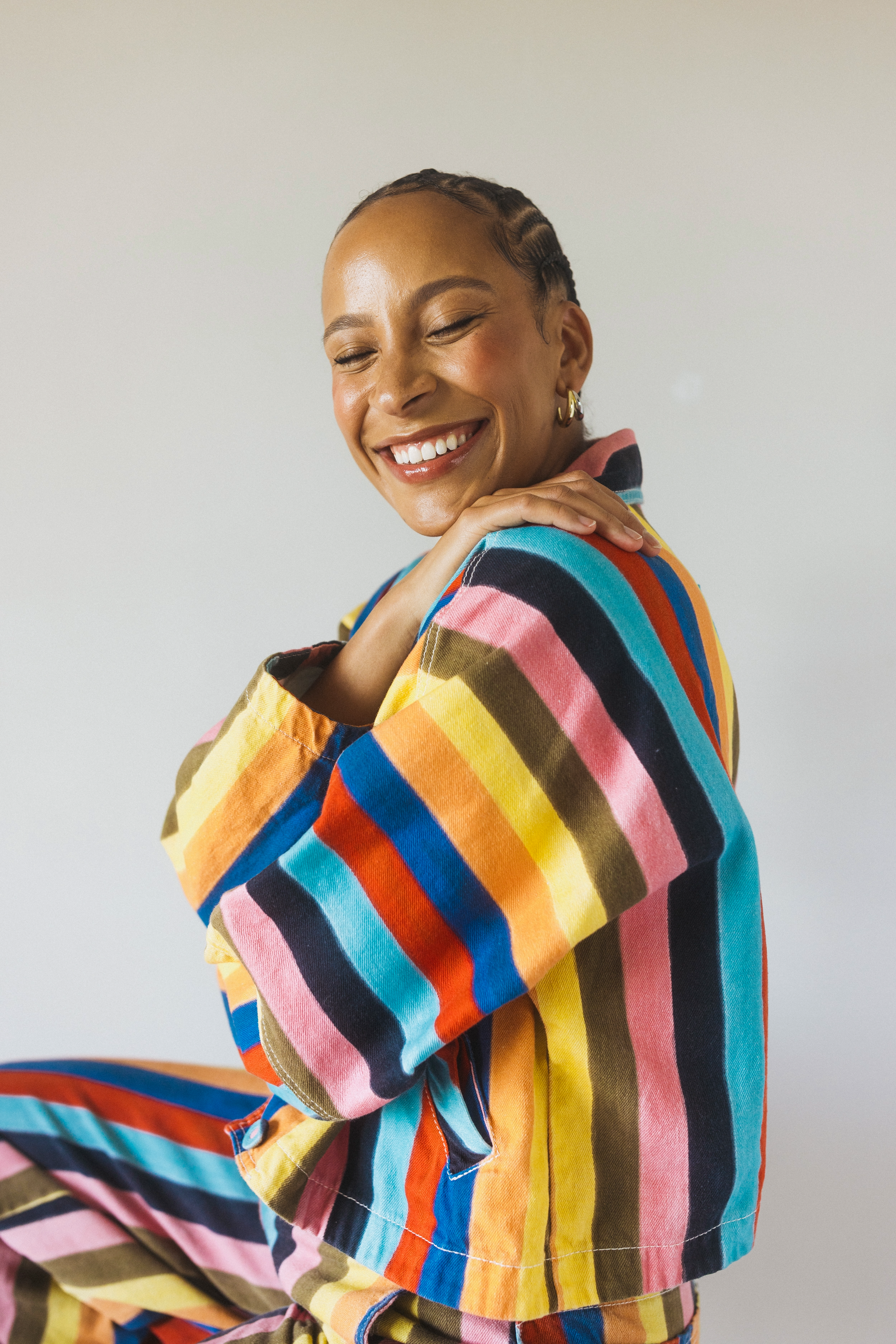 A smiling woman with braided hair and gold hoop earrings wearing a colorful striped jacket posing against a plain neutral background.