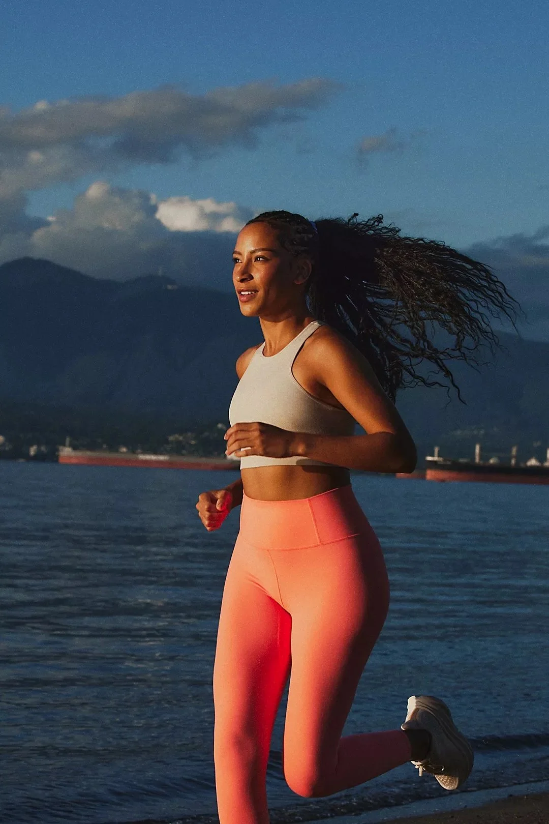 A woman running along a waterfront during sunset, with mountains and water in the background.