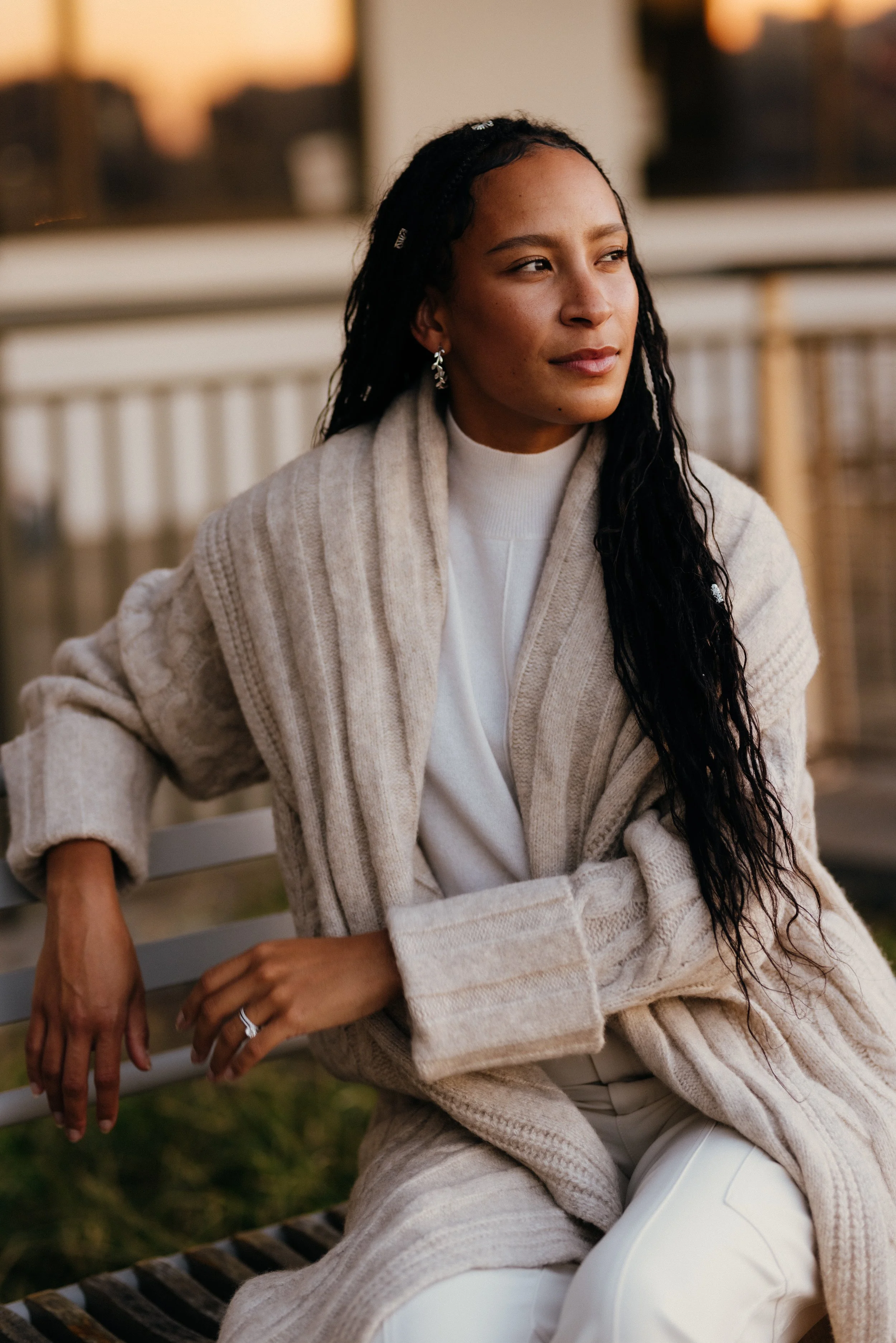 A woman with long, dark, curly hair and earrings sitting on a wooden bench outdoors during sunset, wearing a beige cardigan and white pants.