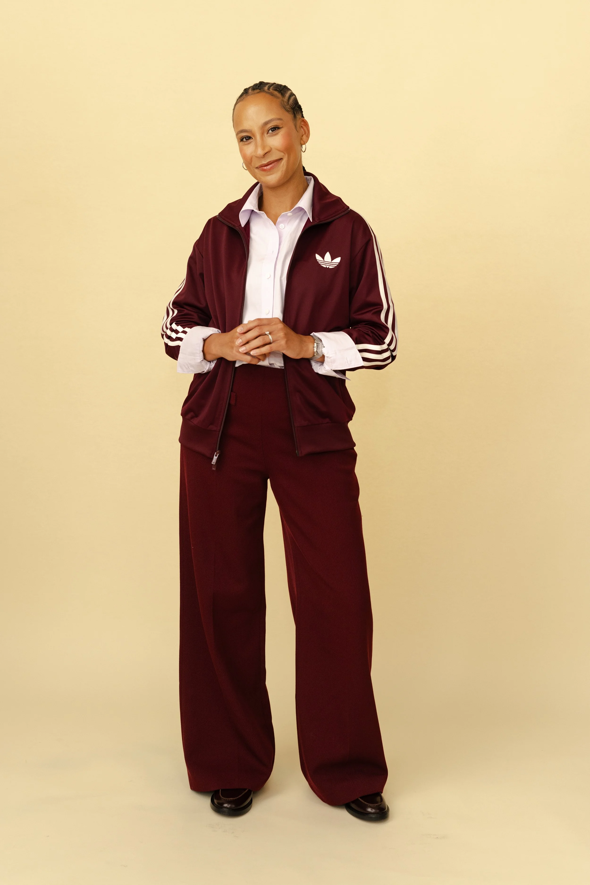 A woman standing against a plain, light-colored background, wearing a white collared shirt, a maroon Adidas track jacket, and wide-leg maroon pants, with black shoes.