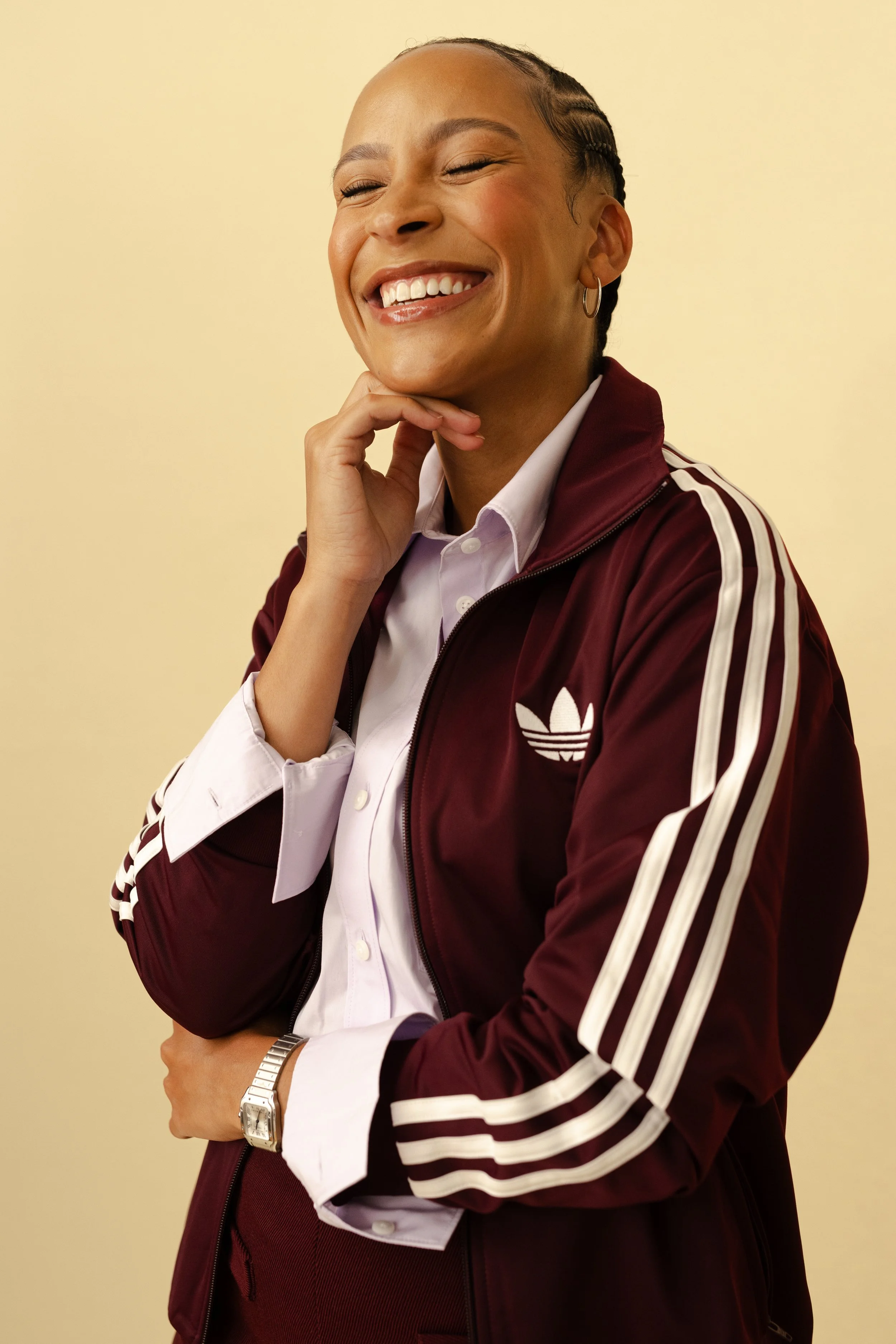 Close-up of a young woman smiling and posing with her hand on her chin, wearing a maroon Adidas jacket with white stripes and a white shirt underneath, against a plain light yellow background.