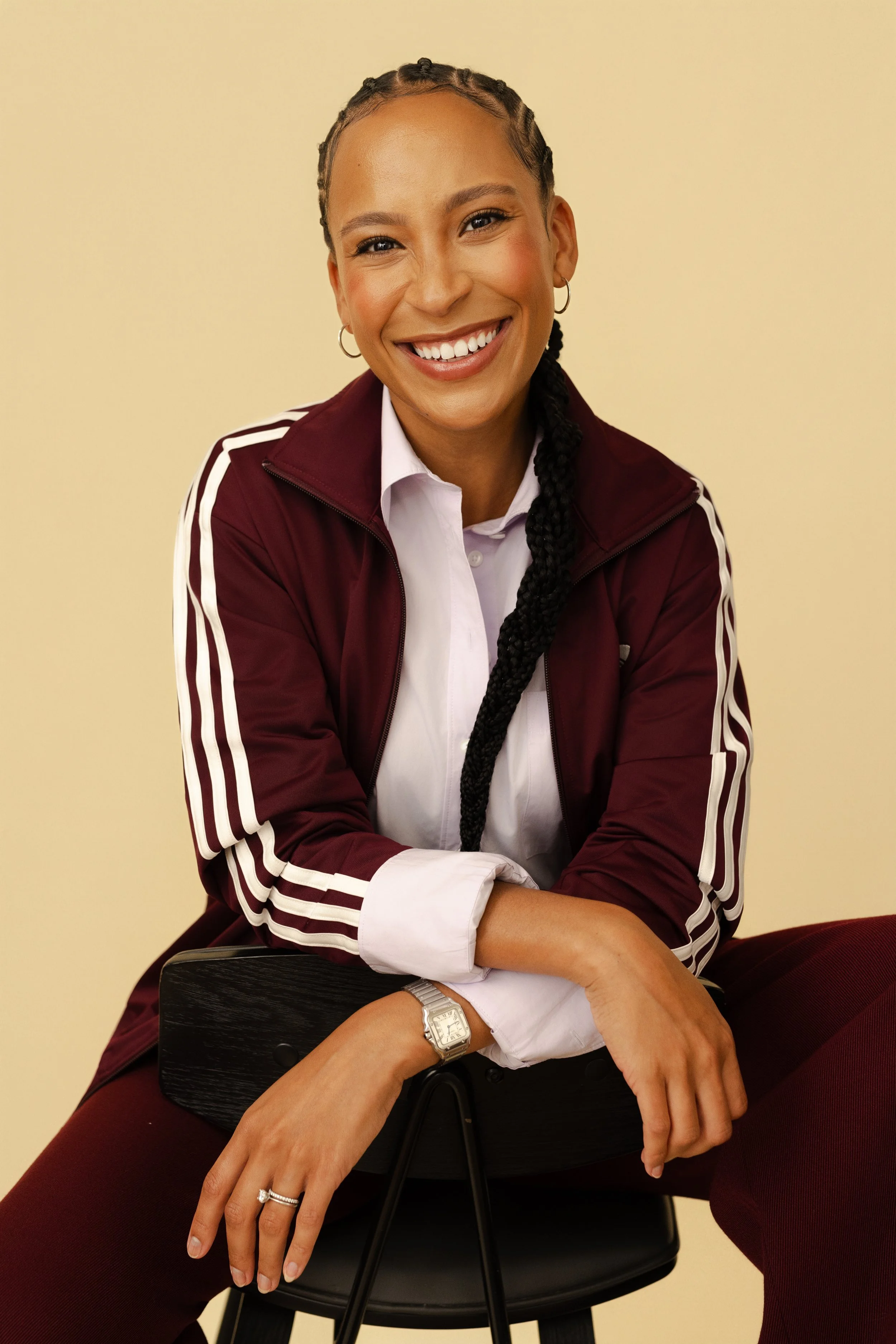 A woman with braids and hoop earrings sitting on a black chair, smiling. She is wearing a white shirt and a maroon zip-up jacket with white stripes on the sleeves. Her right arm is resting on her right knee, and she has a silver watch on her left wrist. The background is a plain light yellow color.