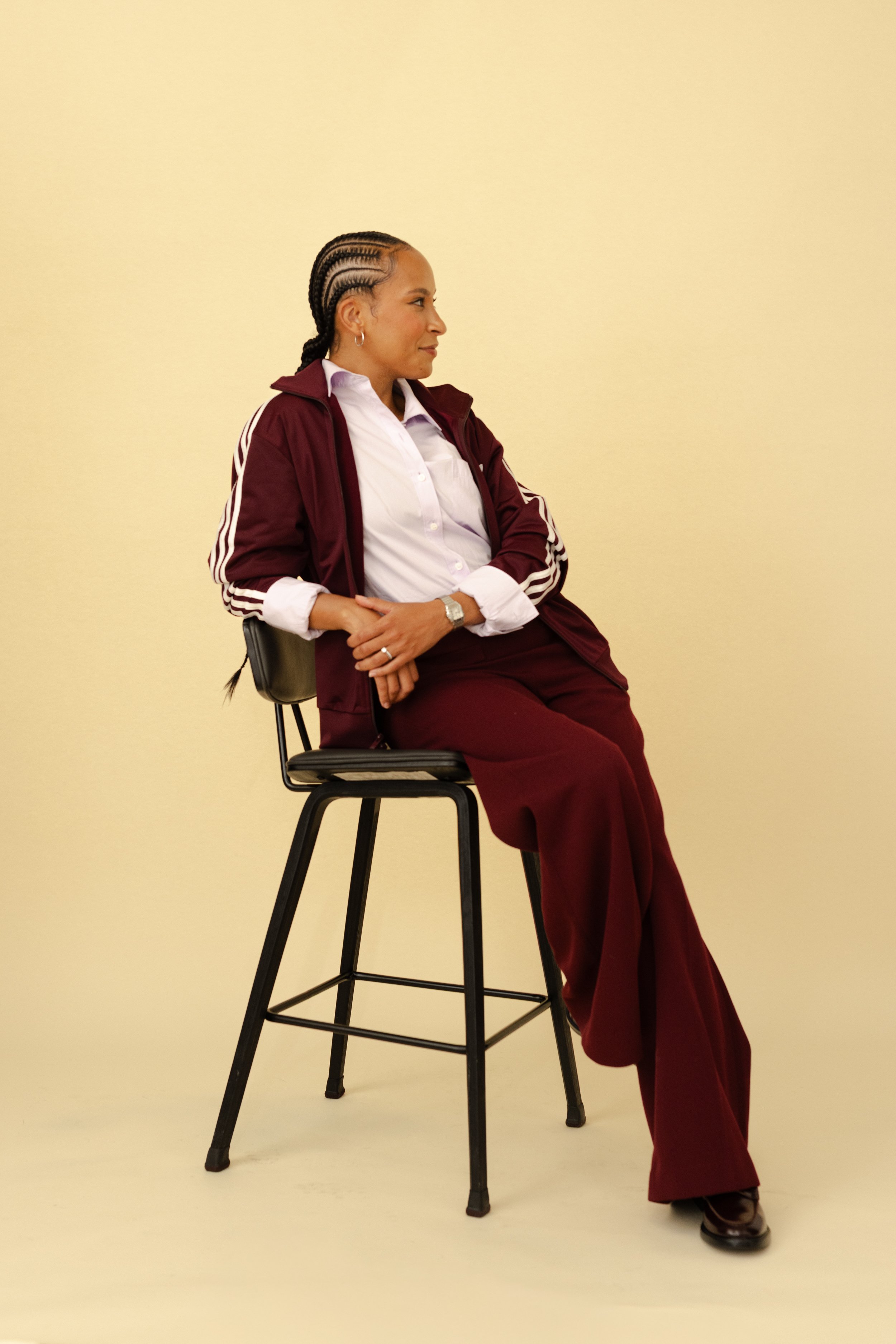 A woman with braided hair, wearing a white shirt and a dark red tracksuit, sitting on a black chair against a yellow background, with her arms crossed and smiling slightly.