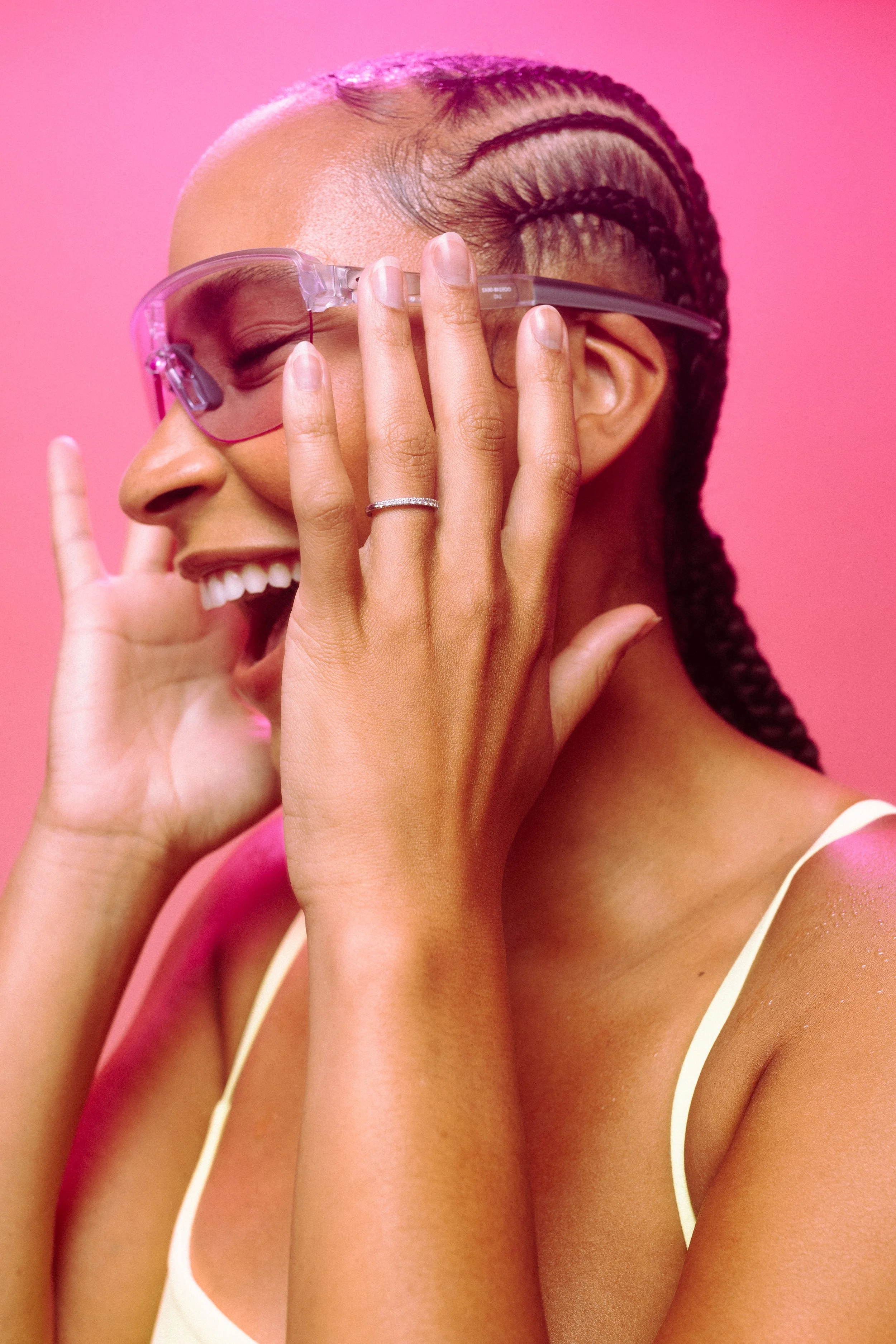 A woman with braided hair wearing pink sunglasses and a yellow spaghetti strap top, smiling against a pink background.