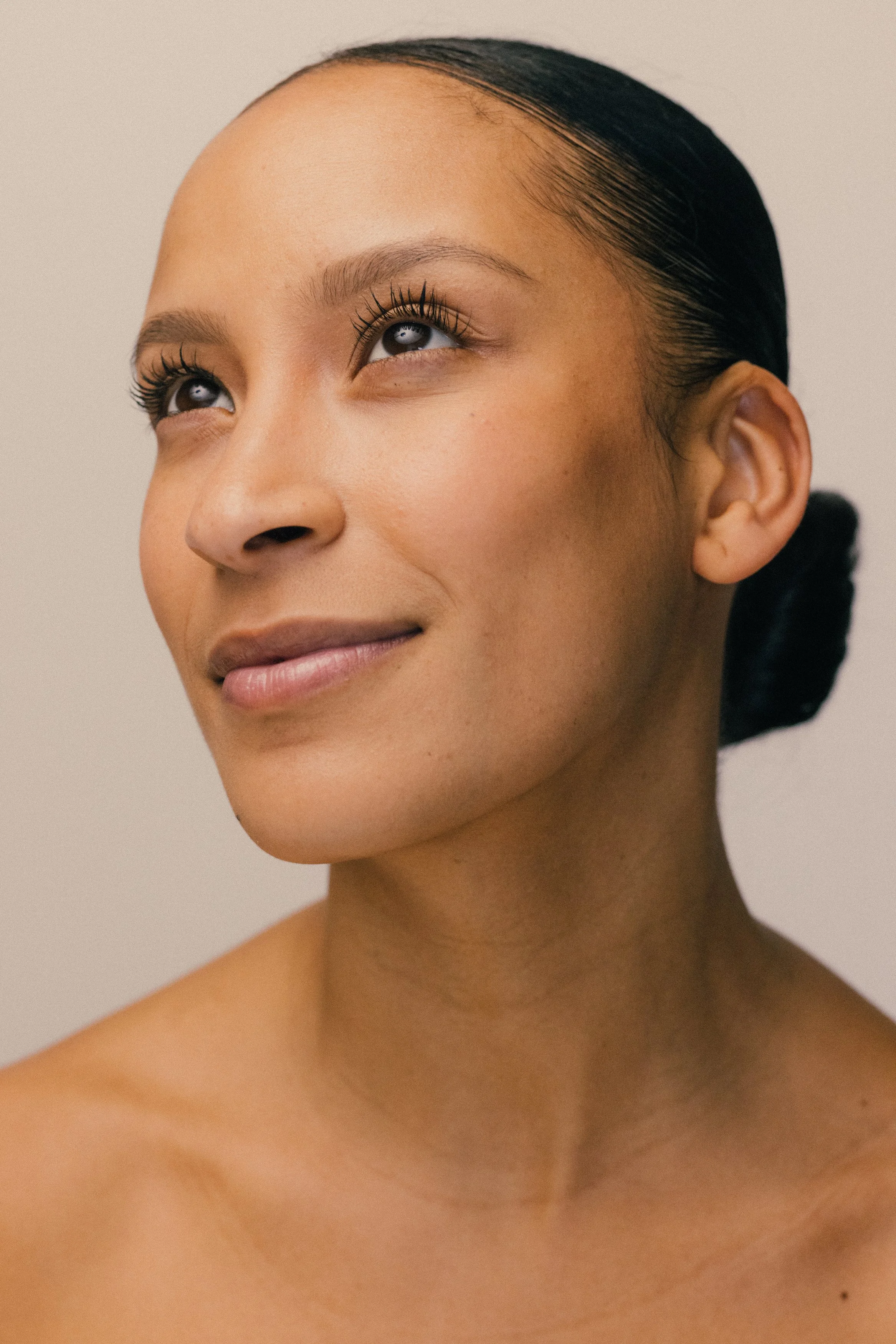 Close-up of a woman with styled dark hair and makeup, looking slightly upwards, against a neutral background.