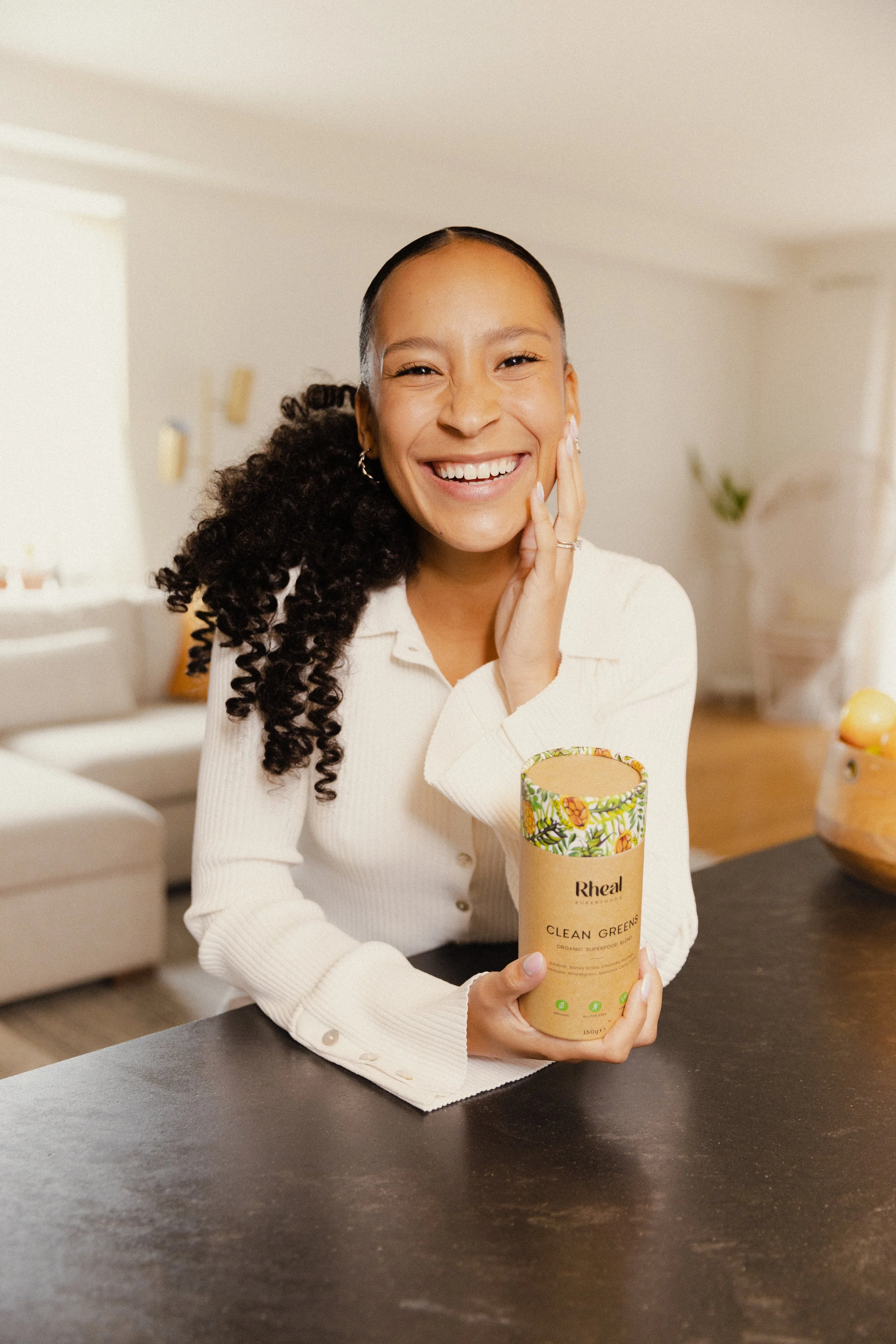 A woman with a curly ponytail and hoop earrings is smiling and touching her face with her right hand. She has light brown skin and is wearing a cream-colored cardigan. She is sitting at a dark kitchen countertop, holding a container of Rheal Superfoo